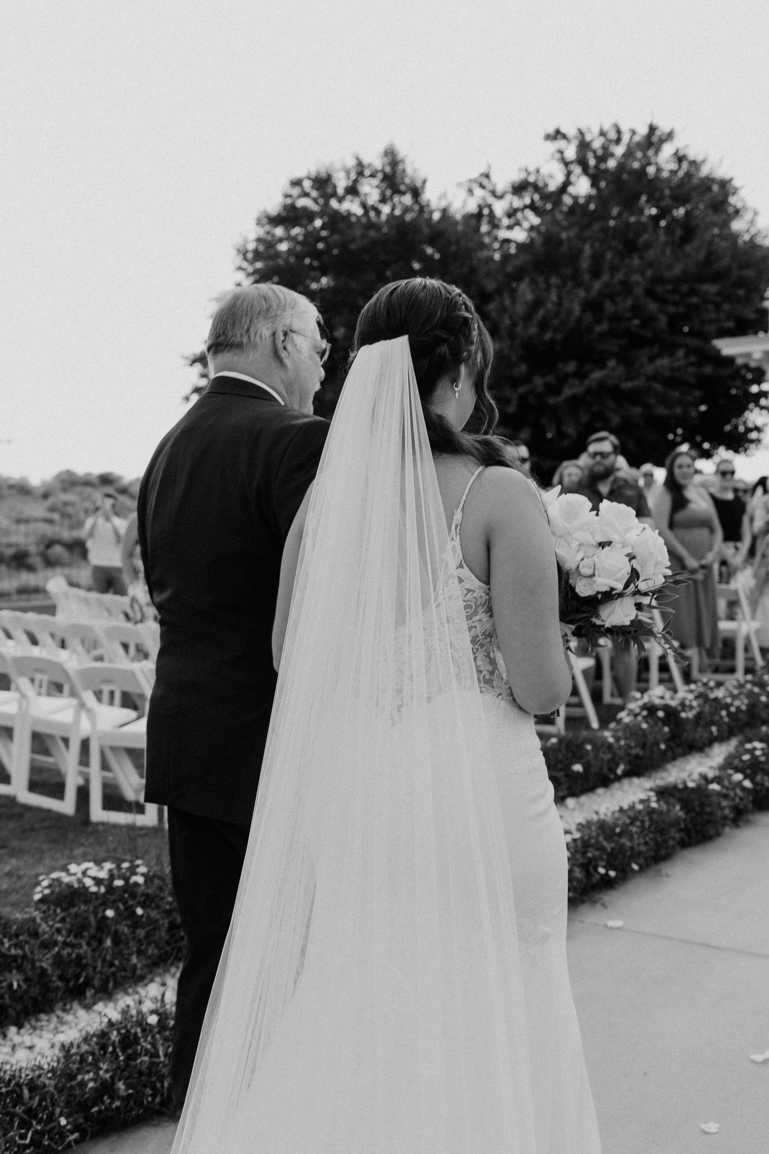 Black and white photo of a bride in a long veil walking with an escort down the aisle at an outdoor garden wedding ceremony.