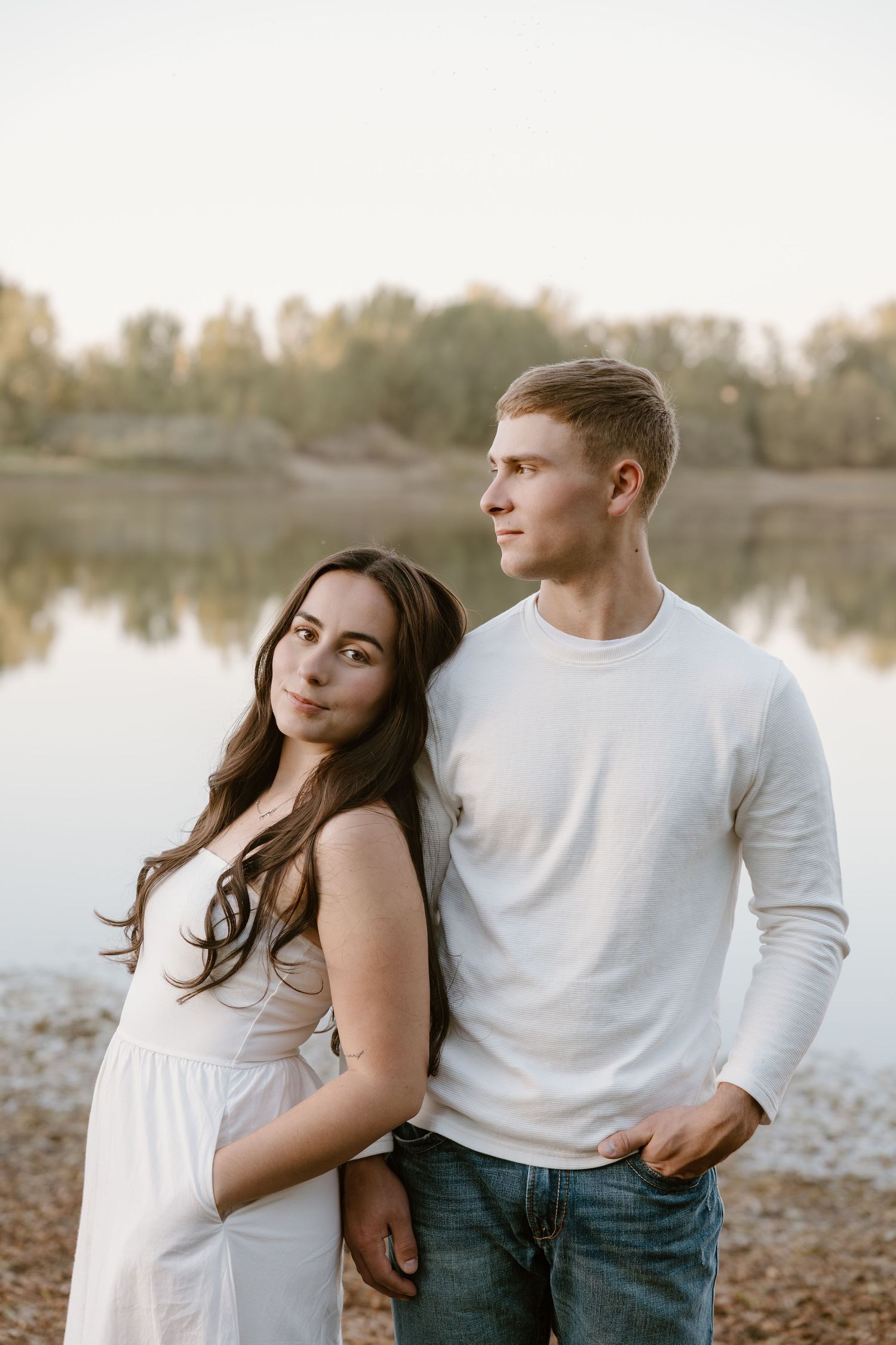 A couple in white shirts stands by a serene lake at sunset, creating a romantic silhouette against the water.