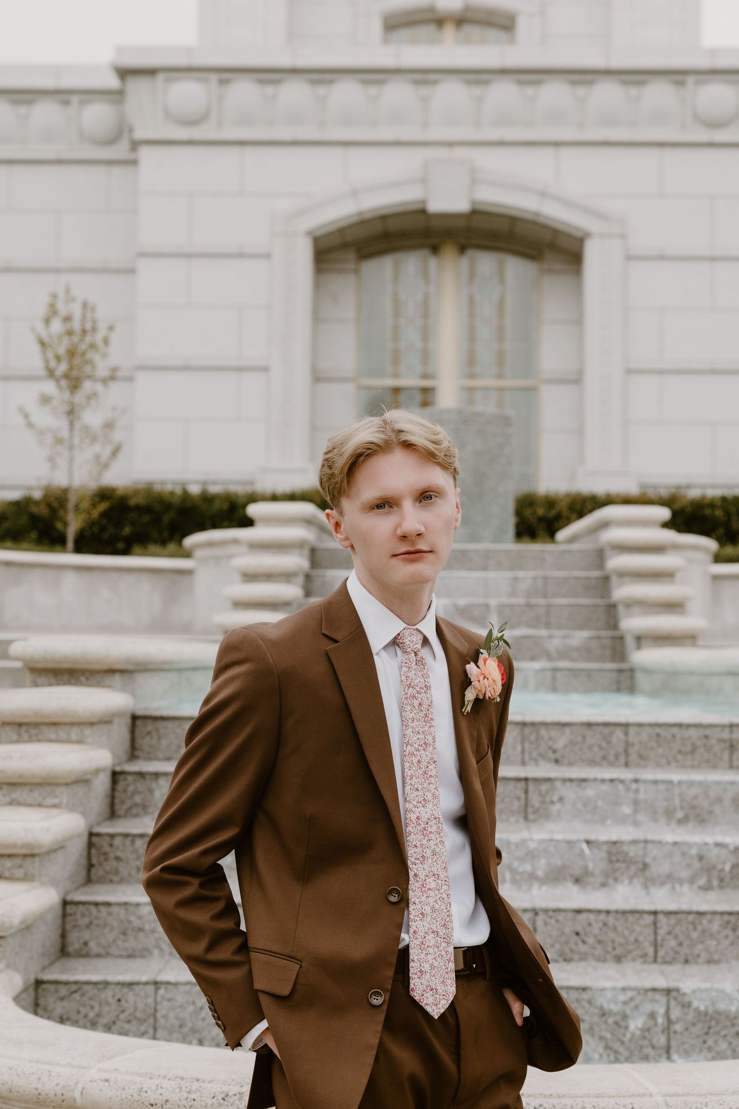A person in a brown suit and floral tie stands on stone steps in front of a white building with classical architecture.
