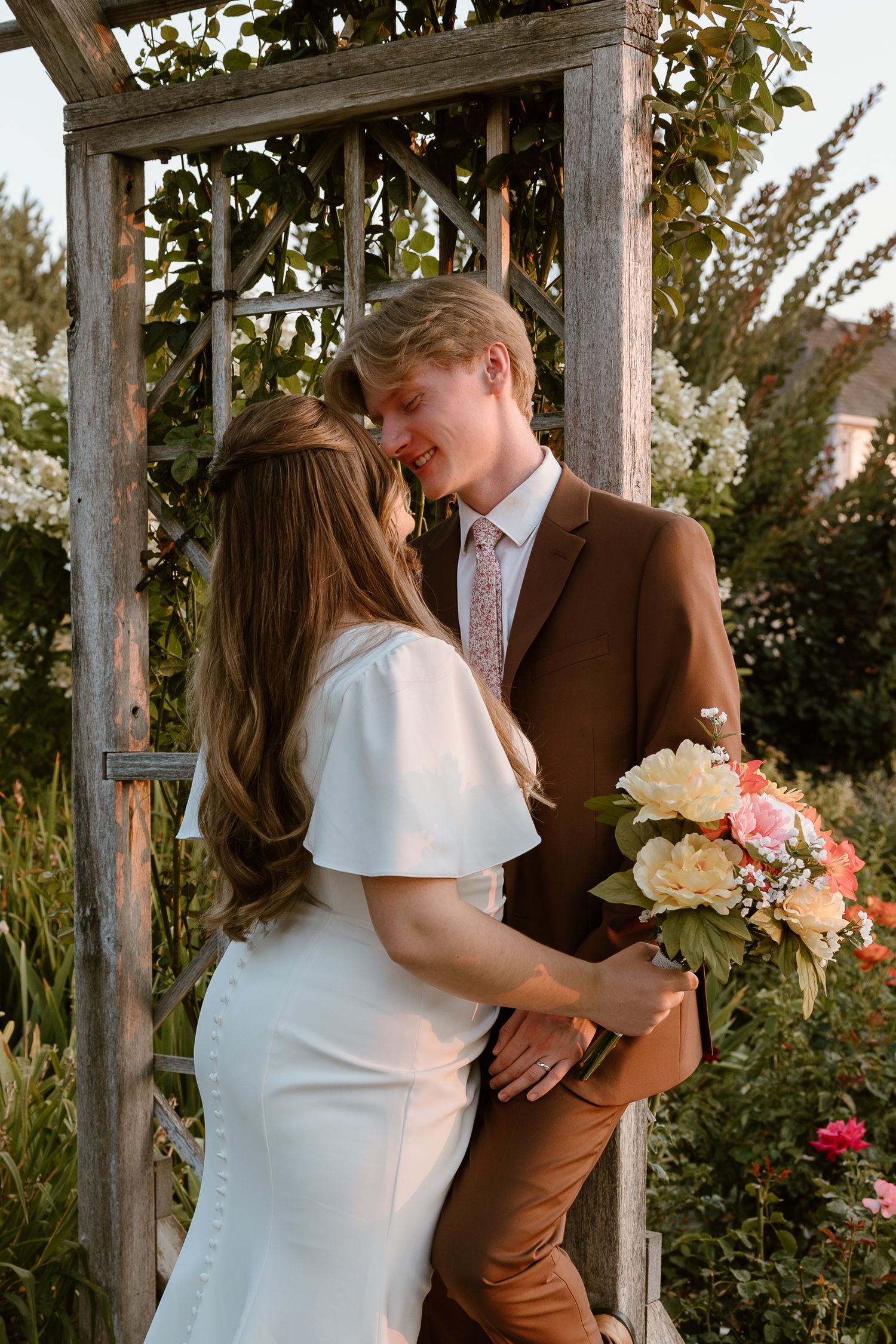 A tender wedding moment captured at sunset as a couple embraces under a rustic wooden arbor with flowers.