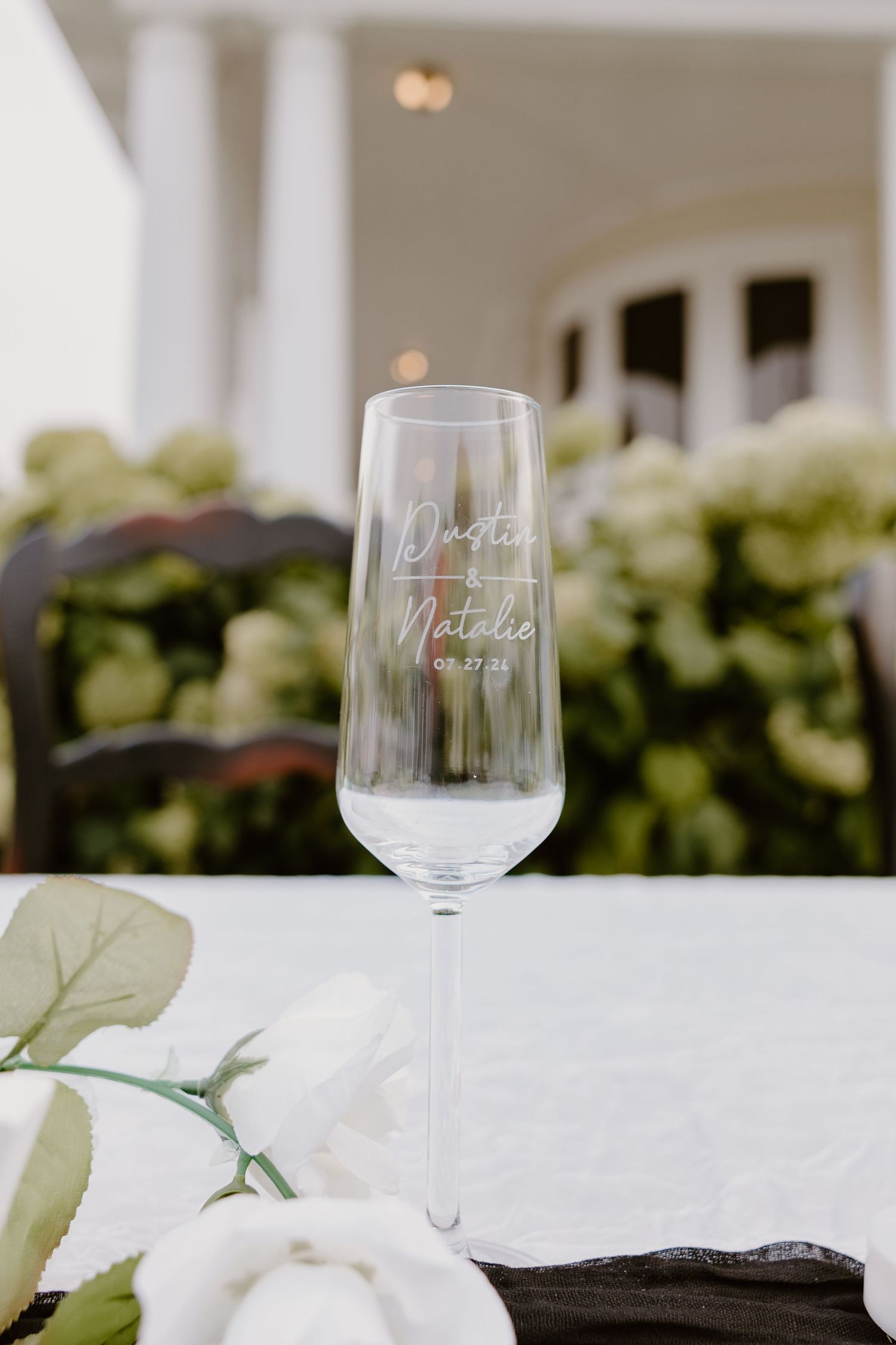 Elegant wine glass with etched lettering sits on a white tablecloth with soft green floral arrangements in the background.