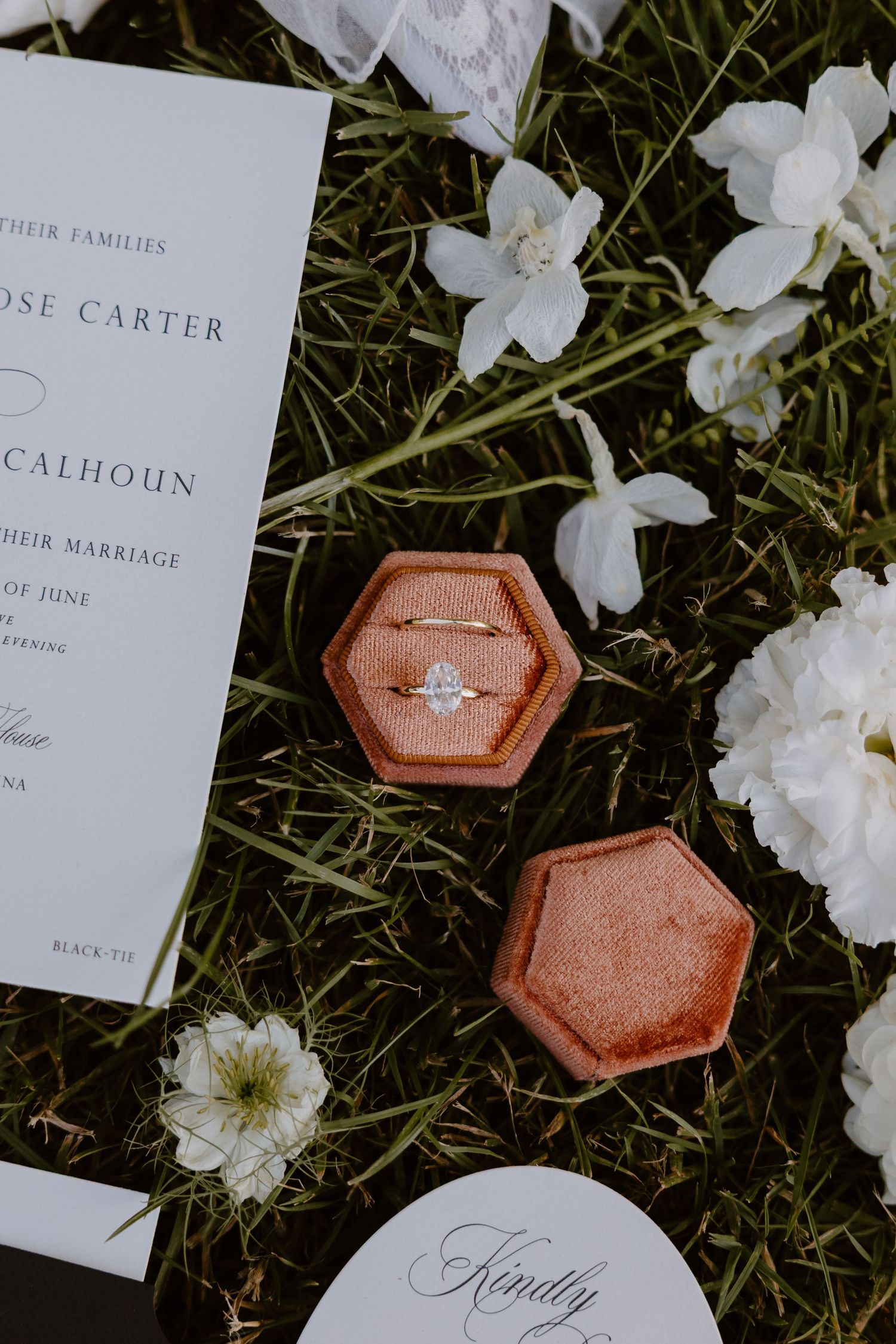 Wedding invitation cards and diamond engagement ring in hexagonal rose gold box displayed on grass with white flowers.