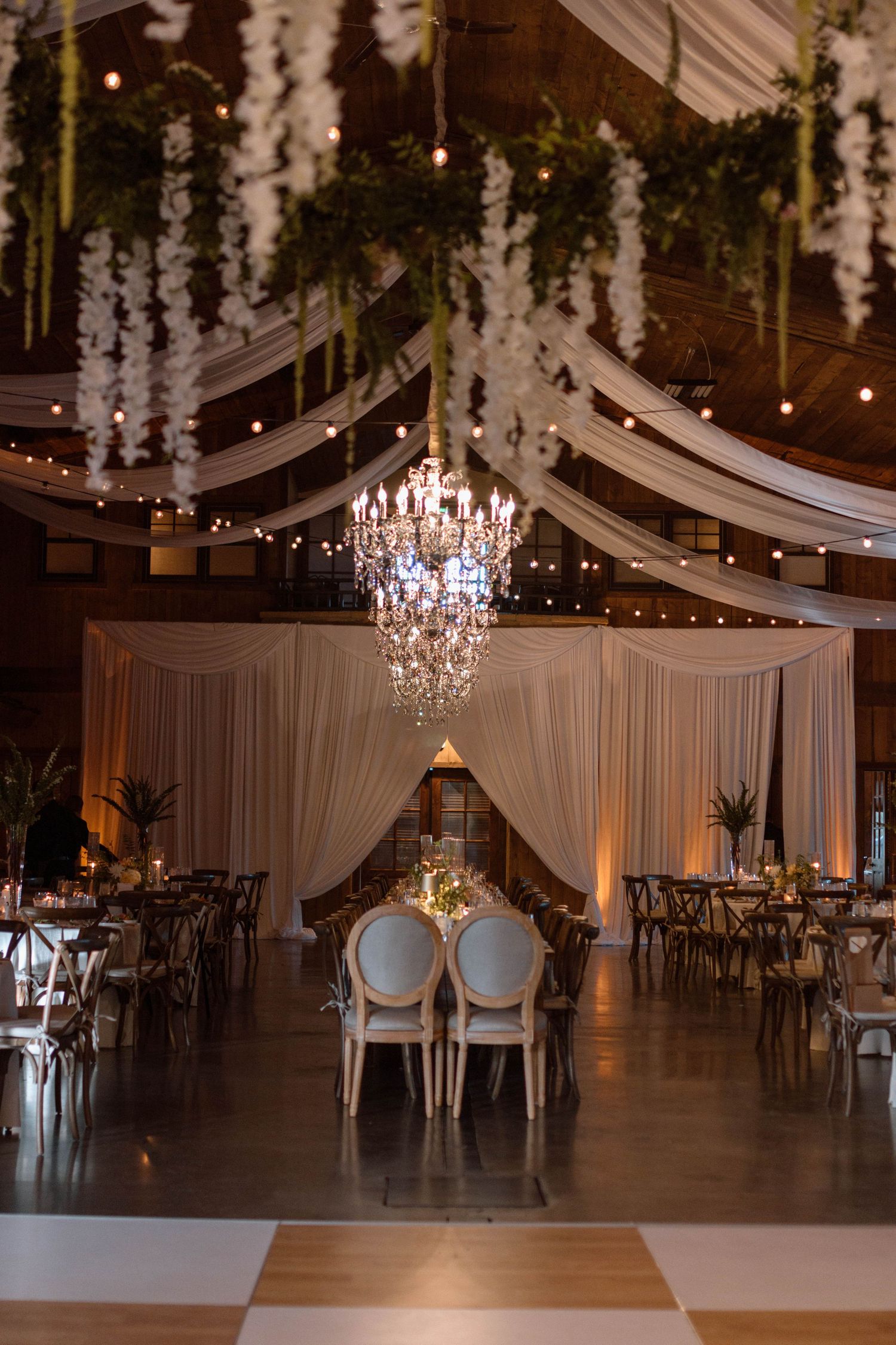 Elegant wedding reception hall with crystal chandelier, draped fabric ceiling, string lights, and floral greenery above dining tables.