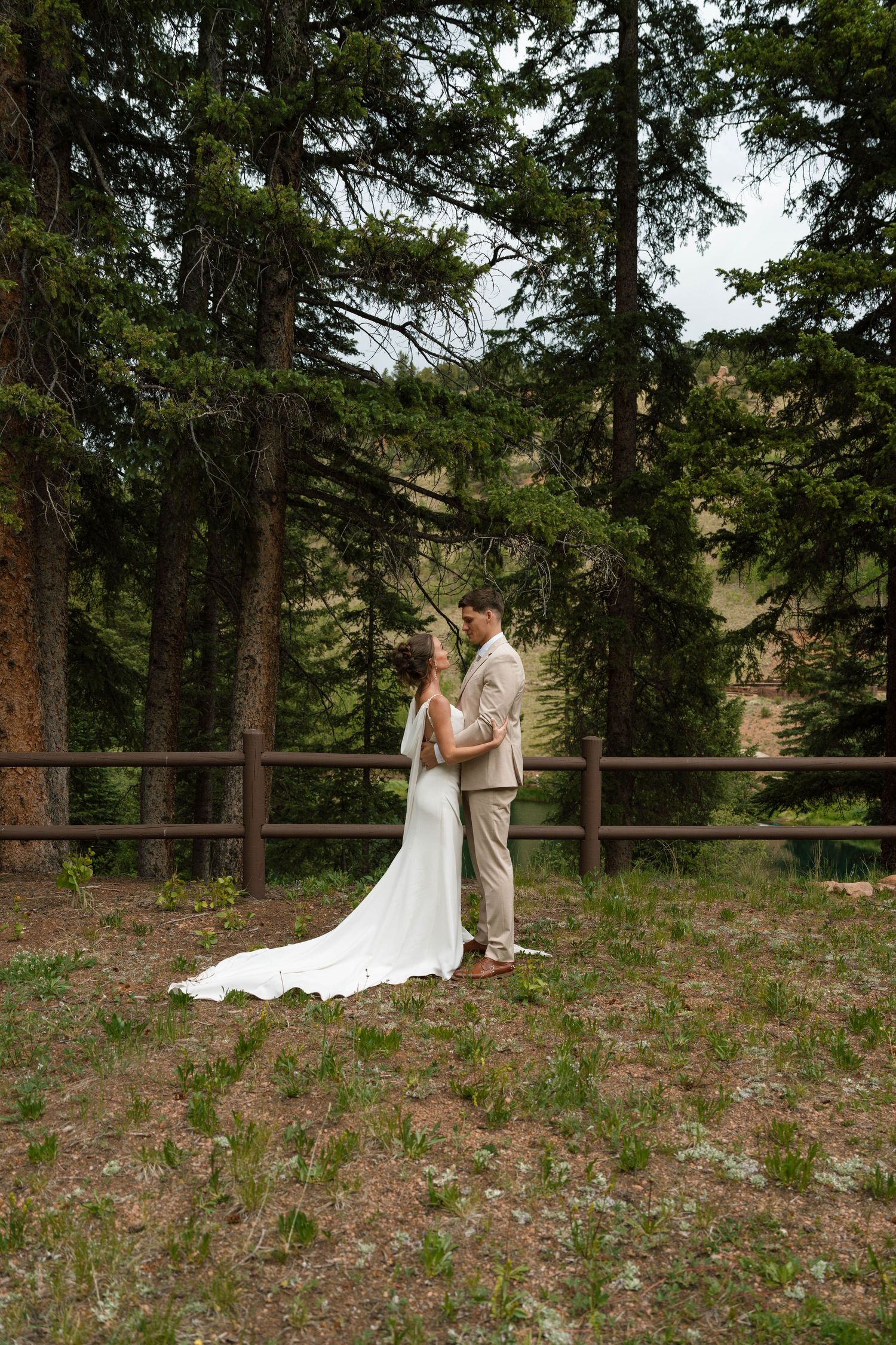 Bride and groom embrace in a forest wedding setting with tall pine trees and rustic wooden fence backdrop.