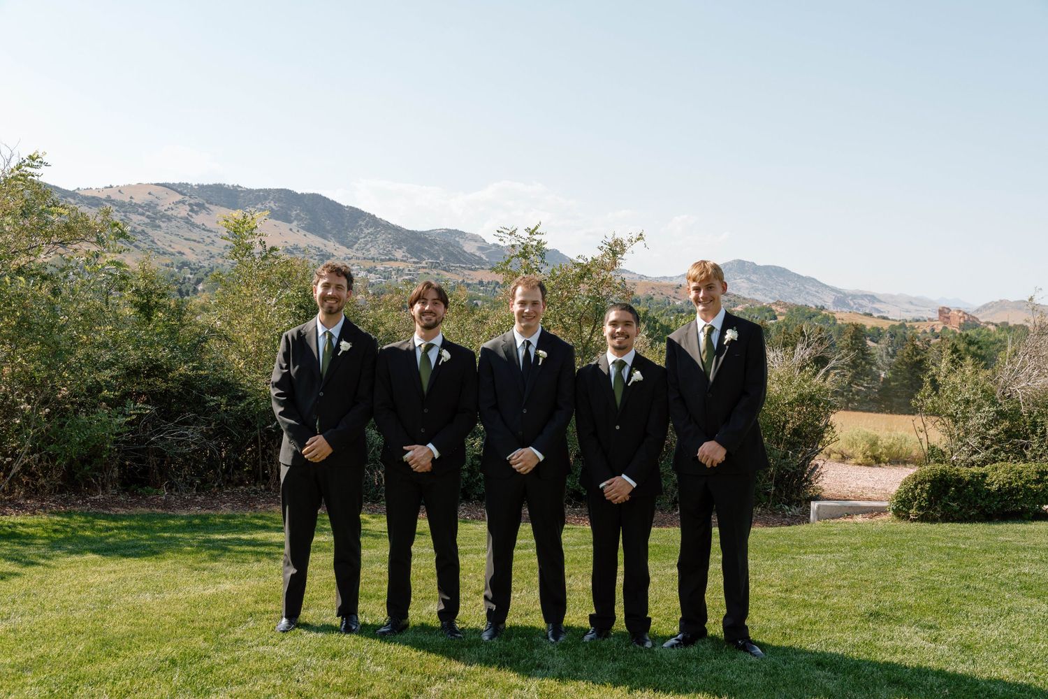 Groomsmen in black suits stand in formation on lawn with mountain backdrop.