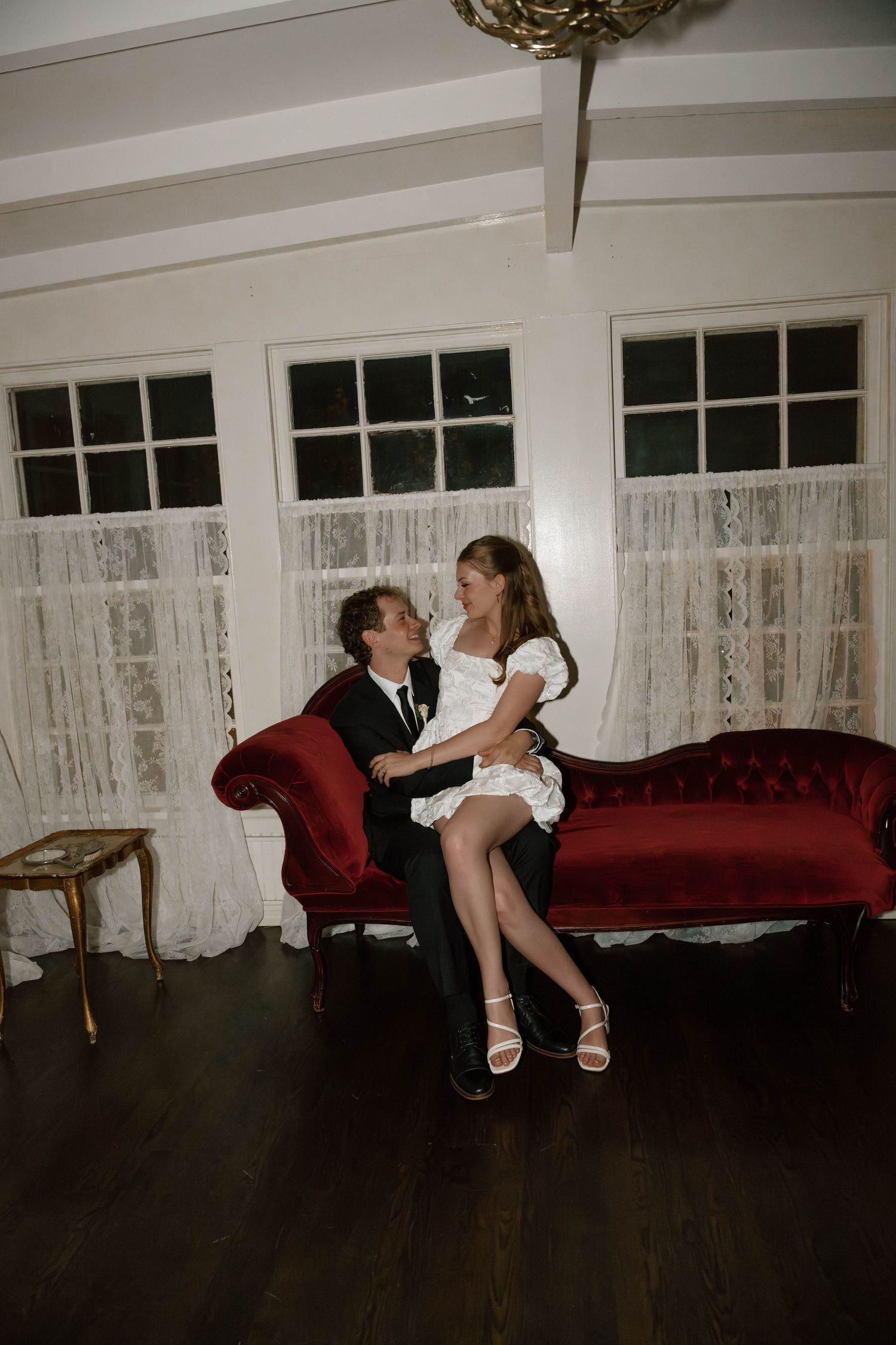 A romantic moment captured on a vintage red velvet couch with lace curtains in the background.
