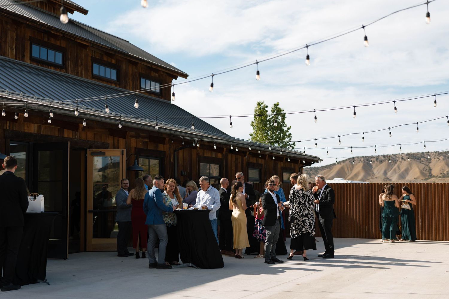 Guests mingle on an outdoor patio at a rustic barn venue with string lights during an evening wedding reception.