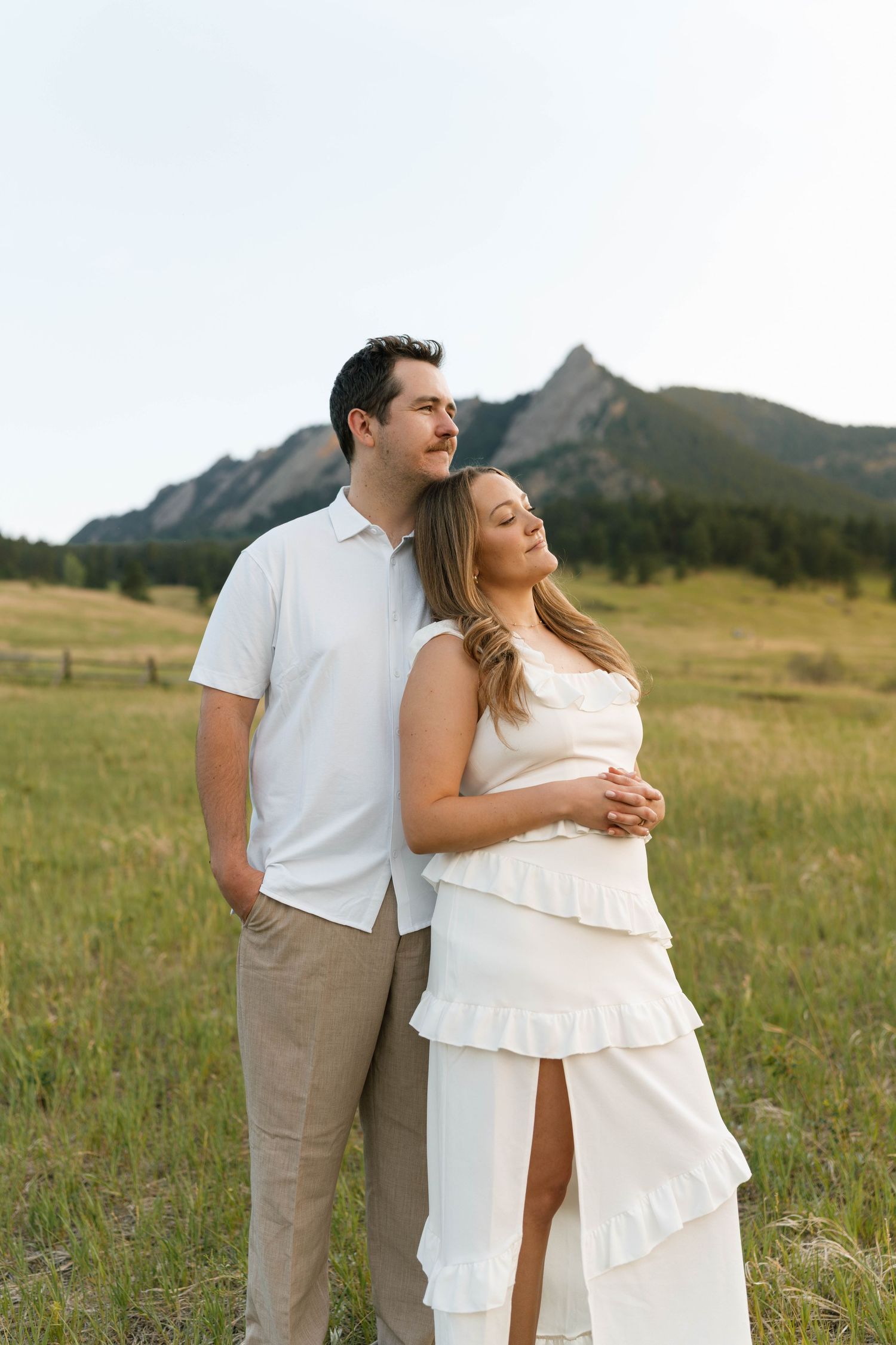 Couple in white outfits embracing in a grassy field with the Flatirons mountains in the background.