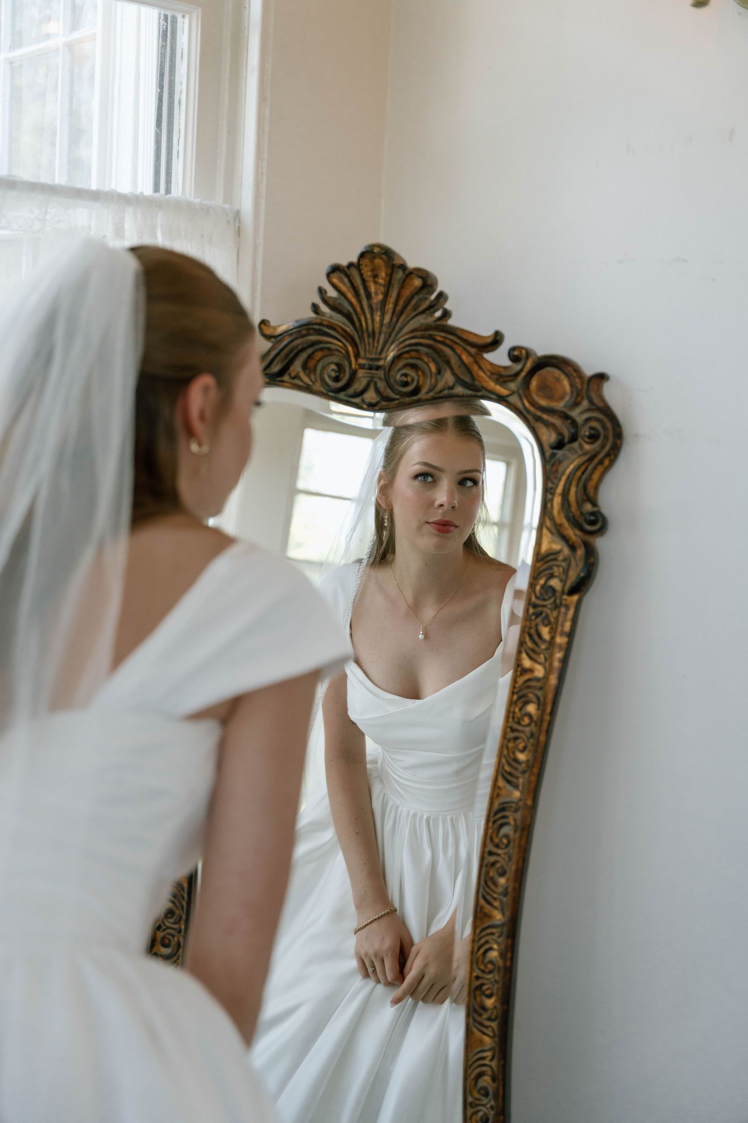 An ornate gold mirror reflects a white dress in a bright minimalist room.