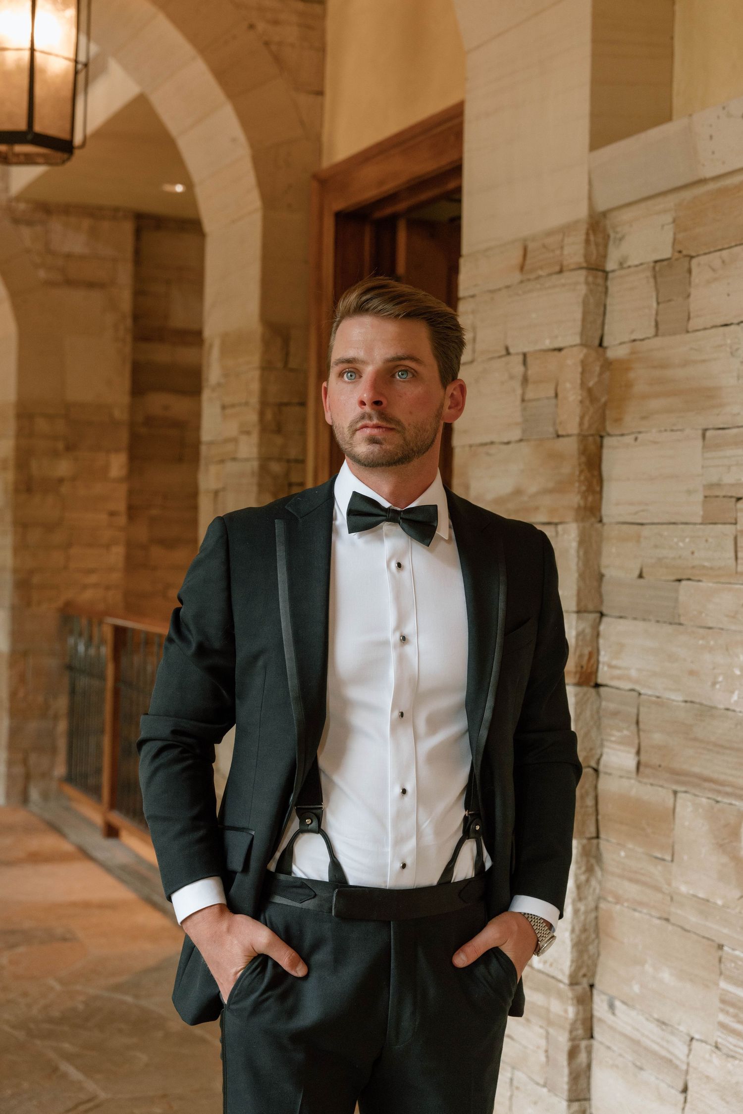 Elegant formal attire with black tuxedo and suspenders against stone wall background.