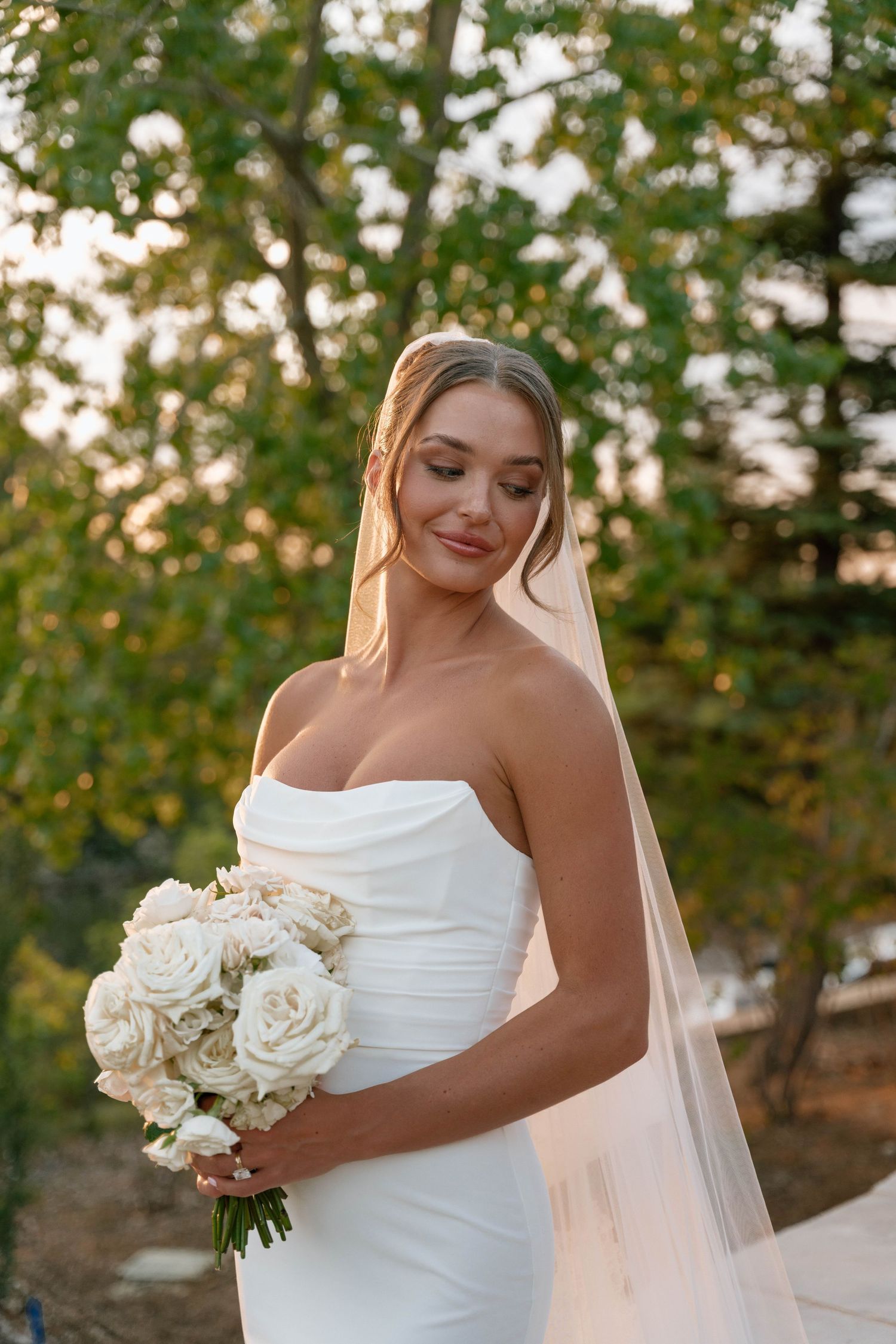 Radiant bride in strapless gown holding white rose bouquet against natural scenery.