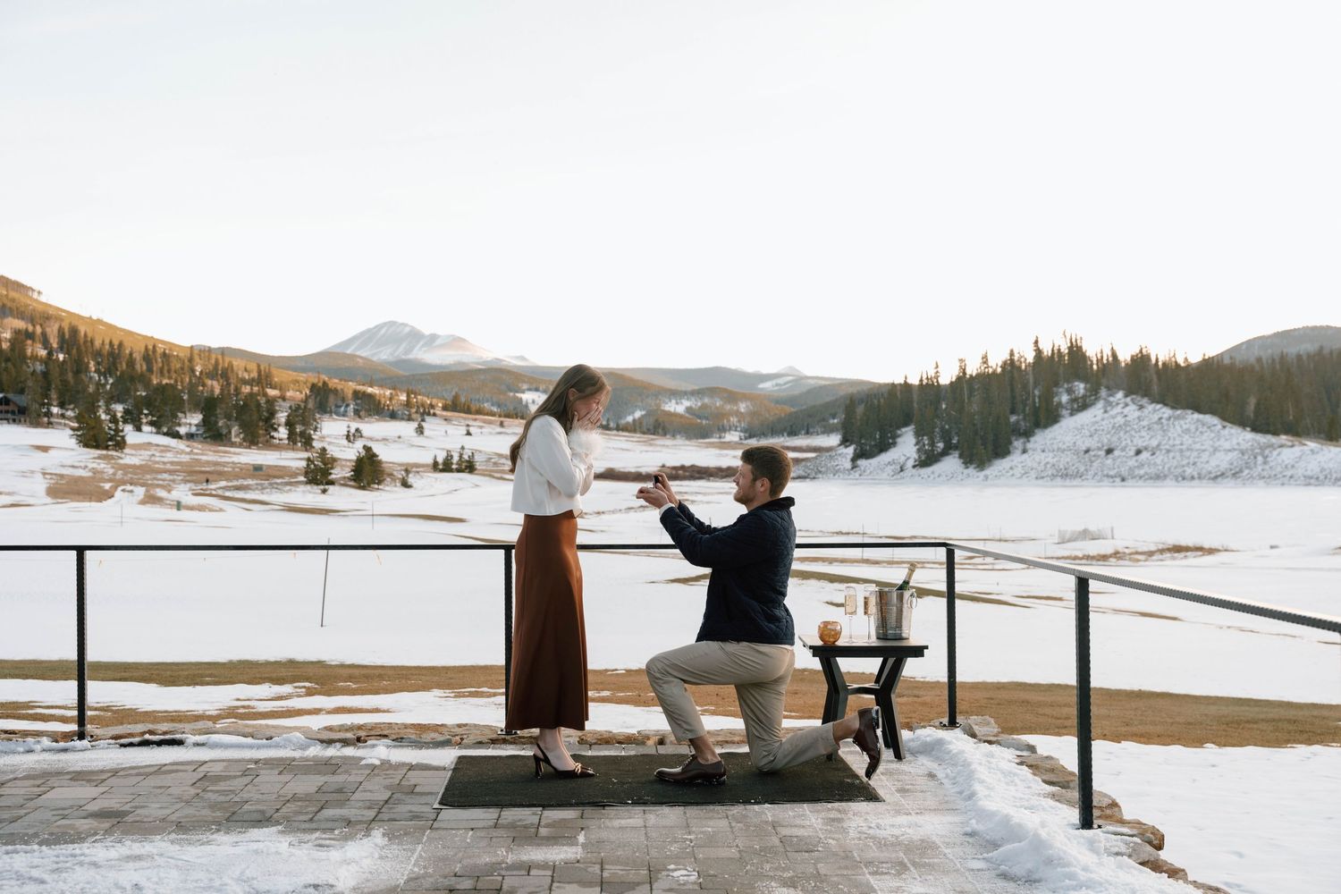 Couple embracing on mountain deck after proposal with champagne and snowy views.