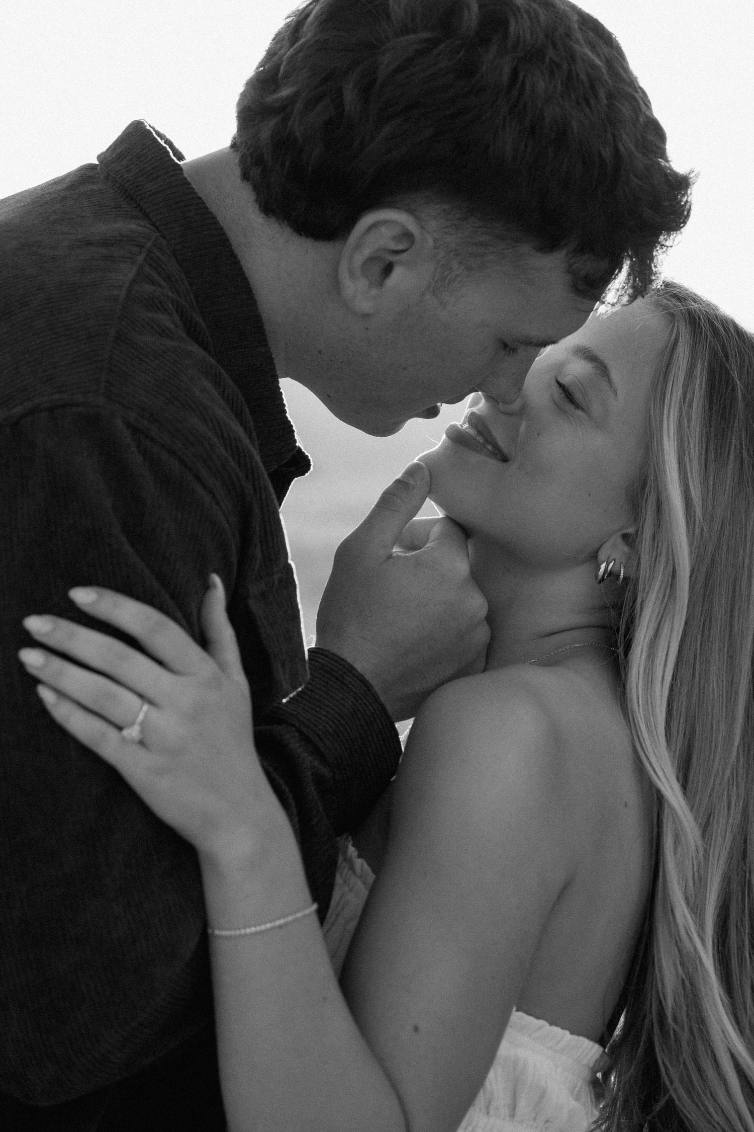 Black and white romantic couple almost kissing, woman wearing engagement ring, tender intimate moment outdoors.