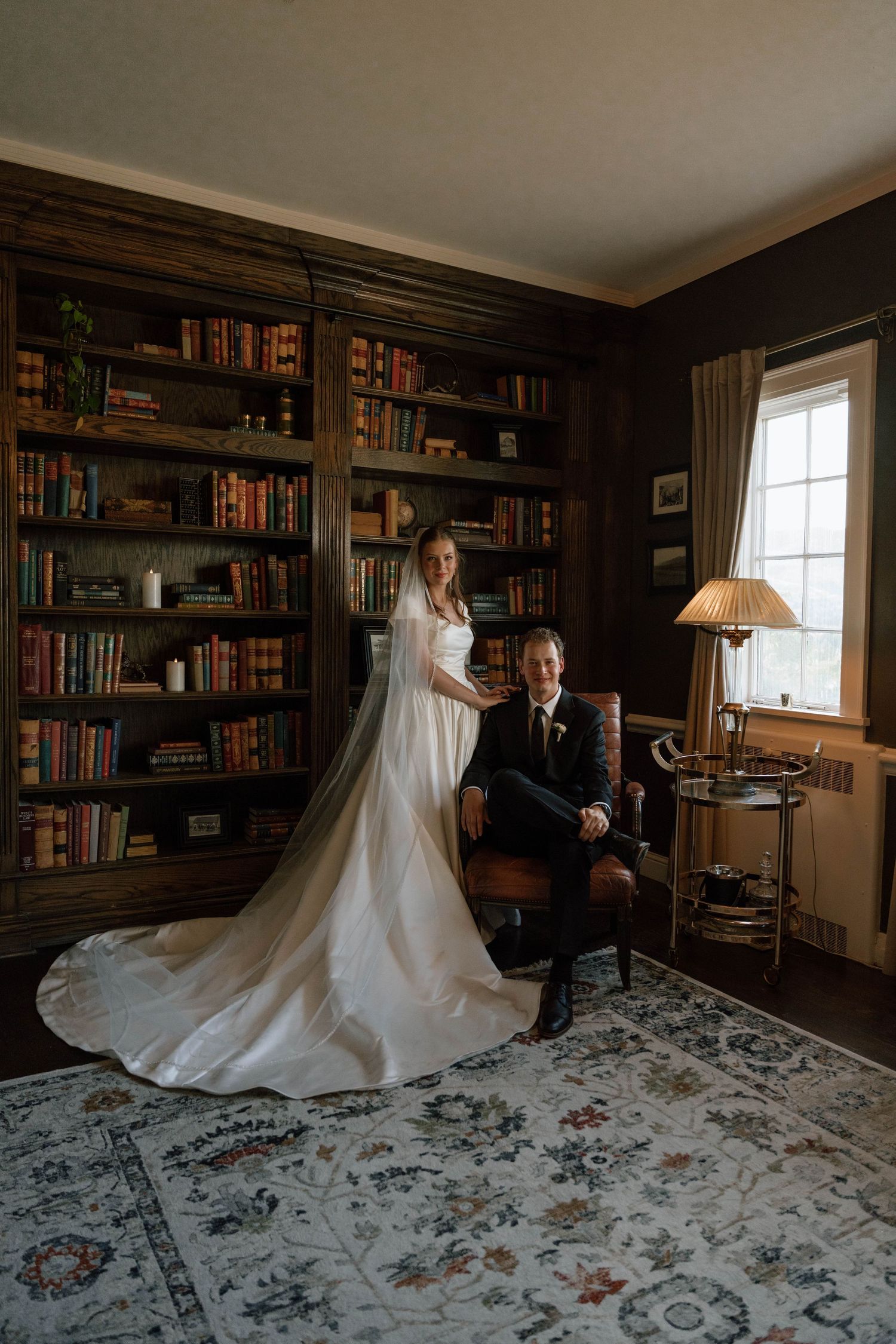 Bride in white gown with veil stands behind groom seated in leather chair in elegant library.