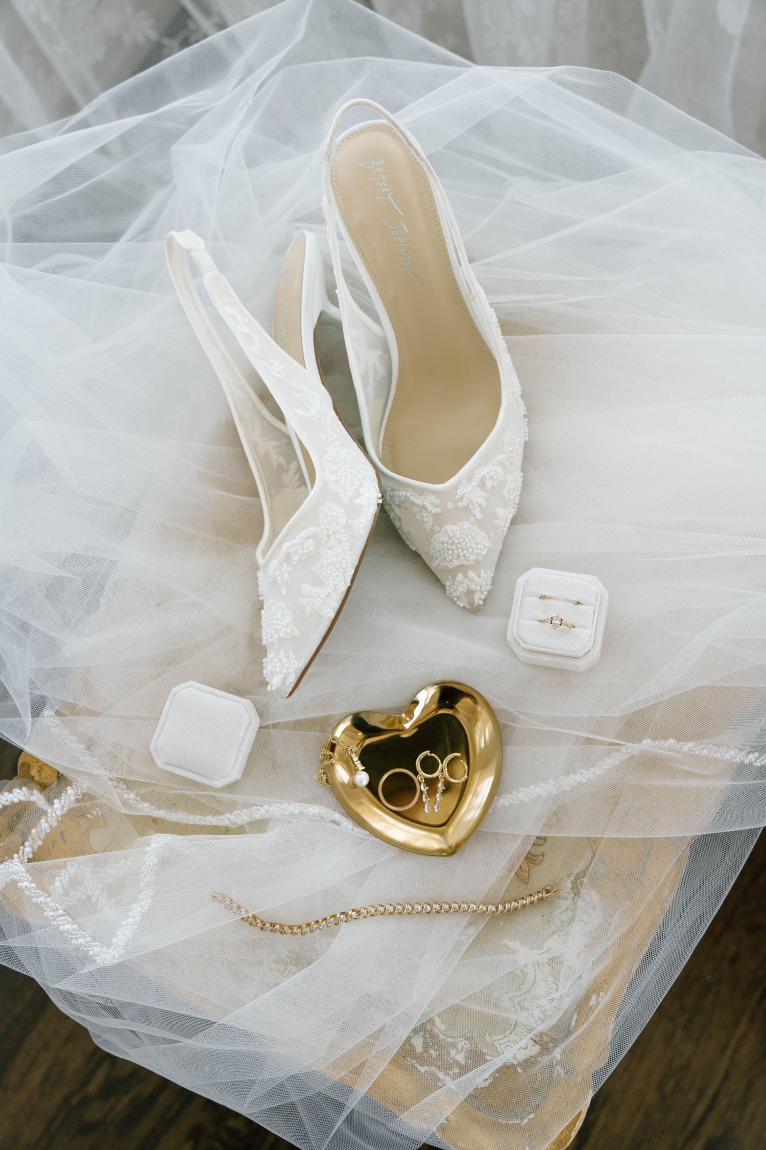 White lace bridal heels displayed with wedding rings in velvet box and gold heart dish on flowing tulle veil.