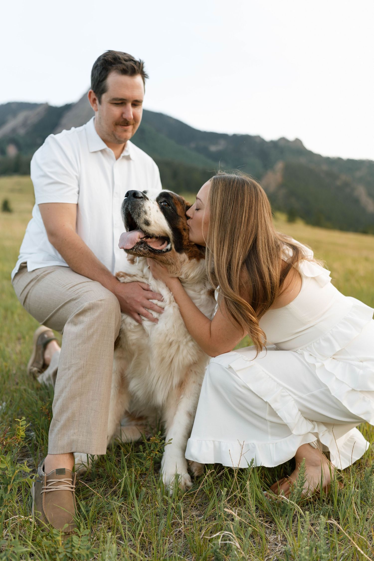 Couple in white outfits kissing their happy dog in a scenic mountain meadow during a golden hour outdoor photo session.
