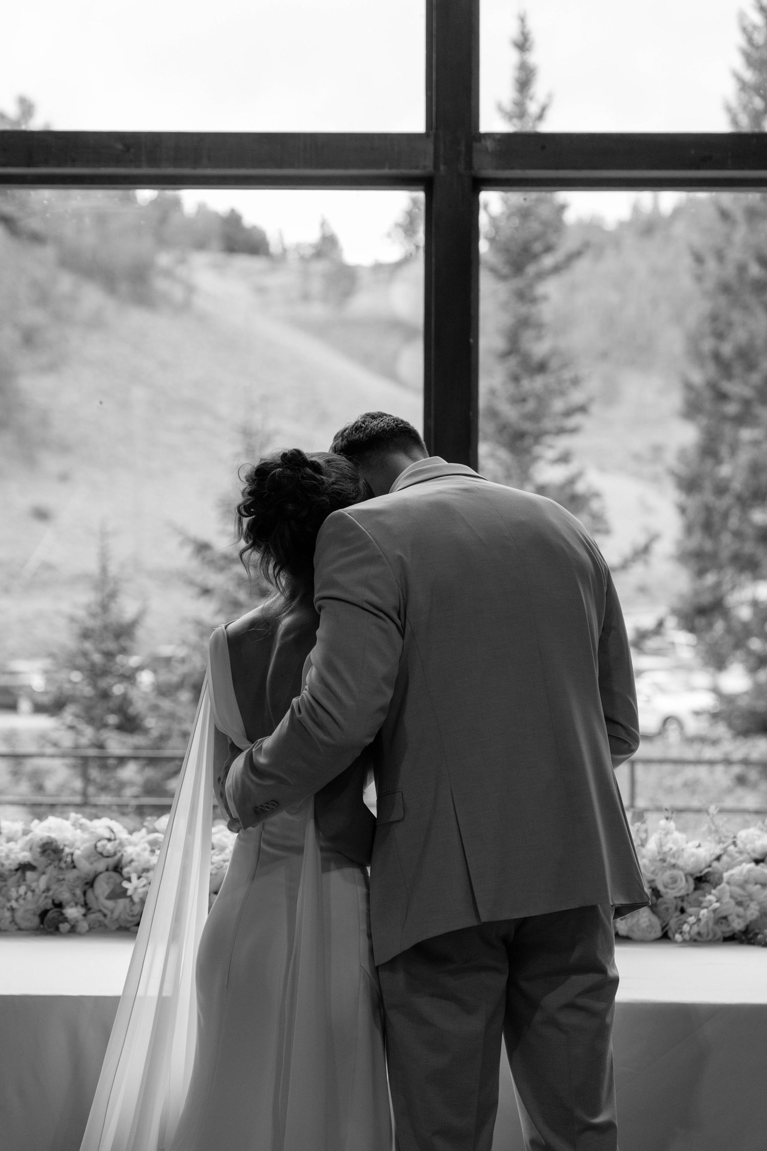 Black and white photo of bride and groom embracing before a large window with mountain views.