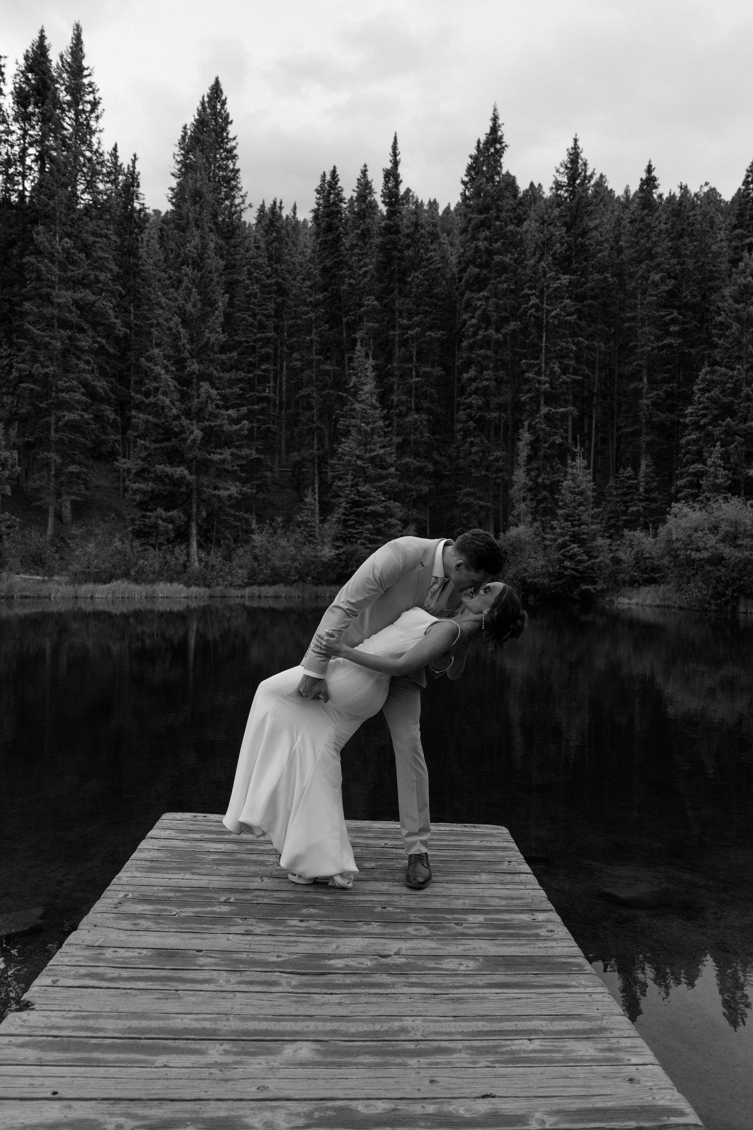 Bride and groom share romantic dip kiss on wooden dock surrounded by pine forest lake.