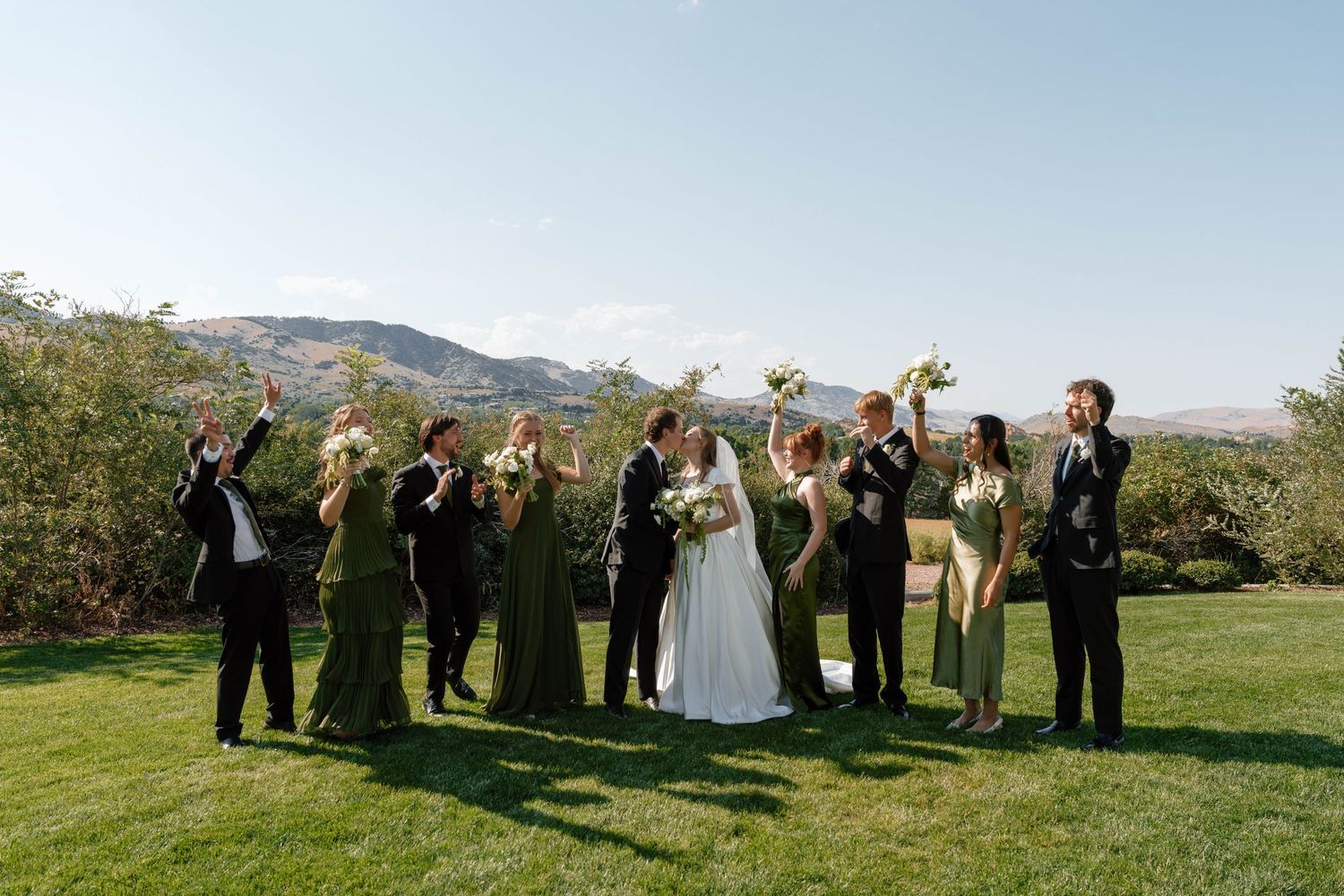 Wedding party poses together on a grassy hill with mountains in the background during an outdoor celebration.