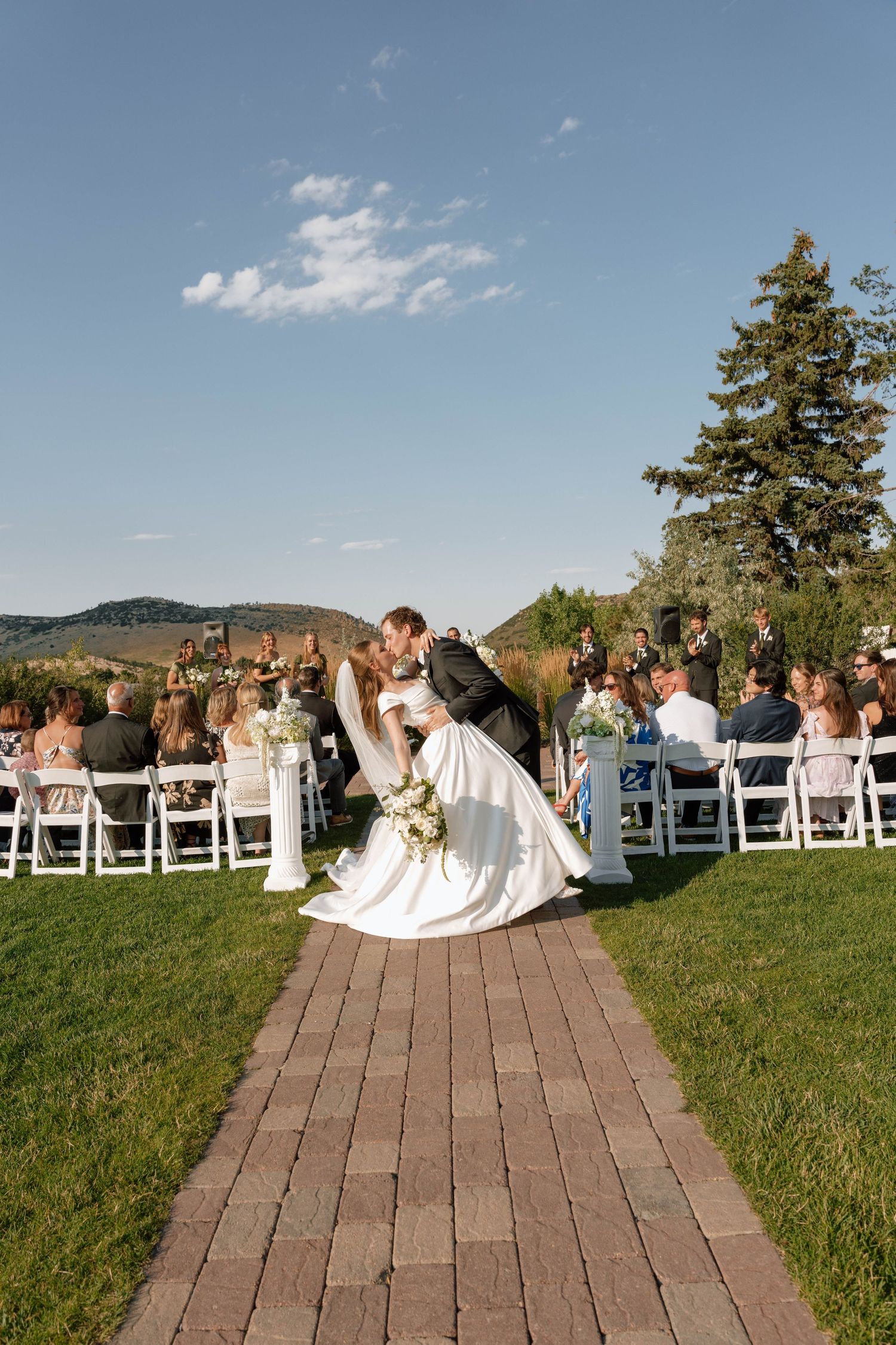 A romantic wedding kiss on a brick pathway with guests and mountains in the background.