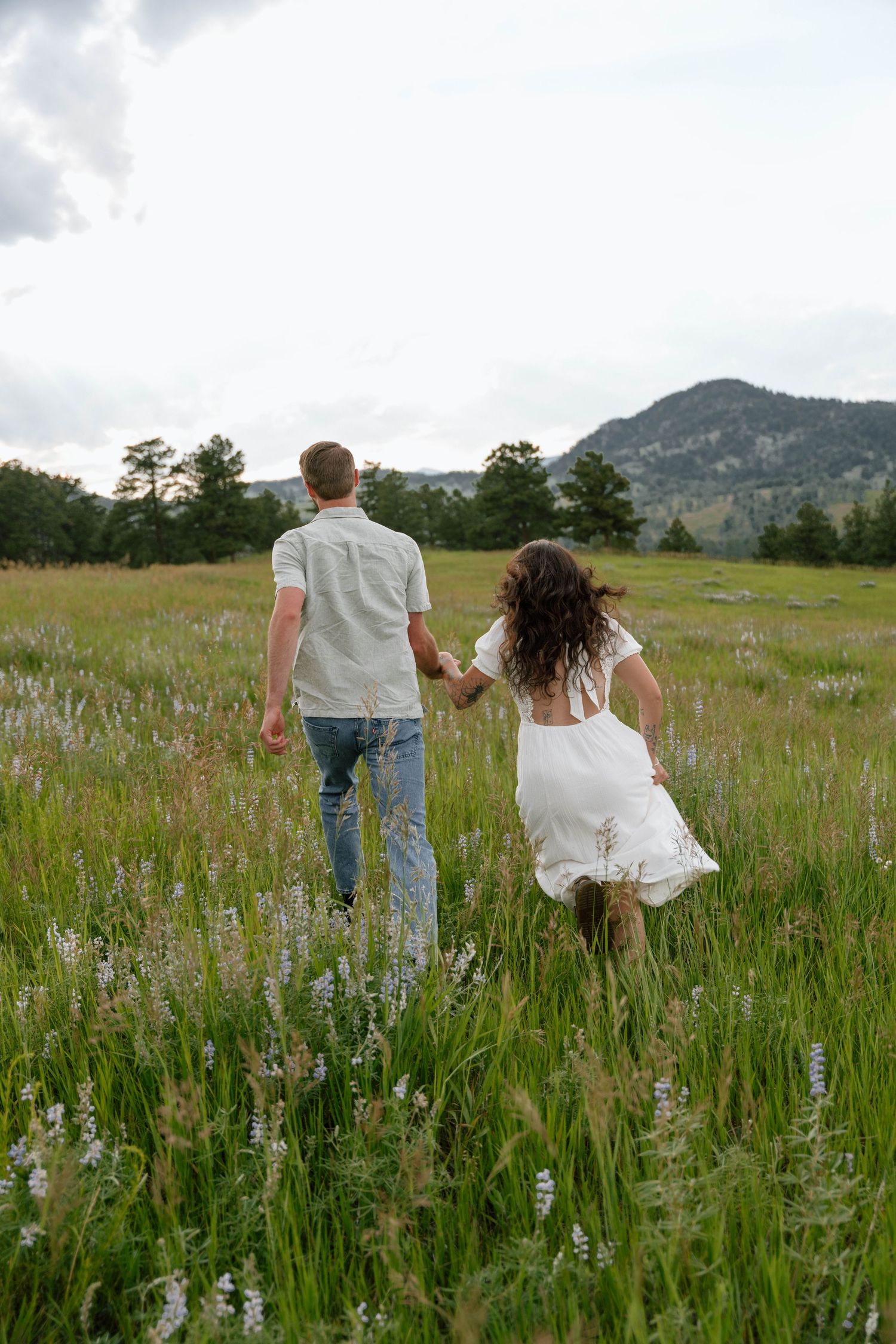 A couple walks through a wildflower meadow with mountains in the background on a summer day.
