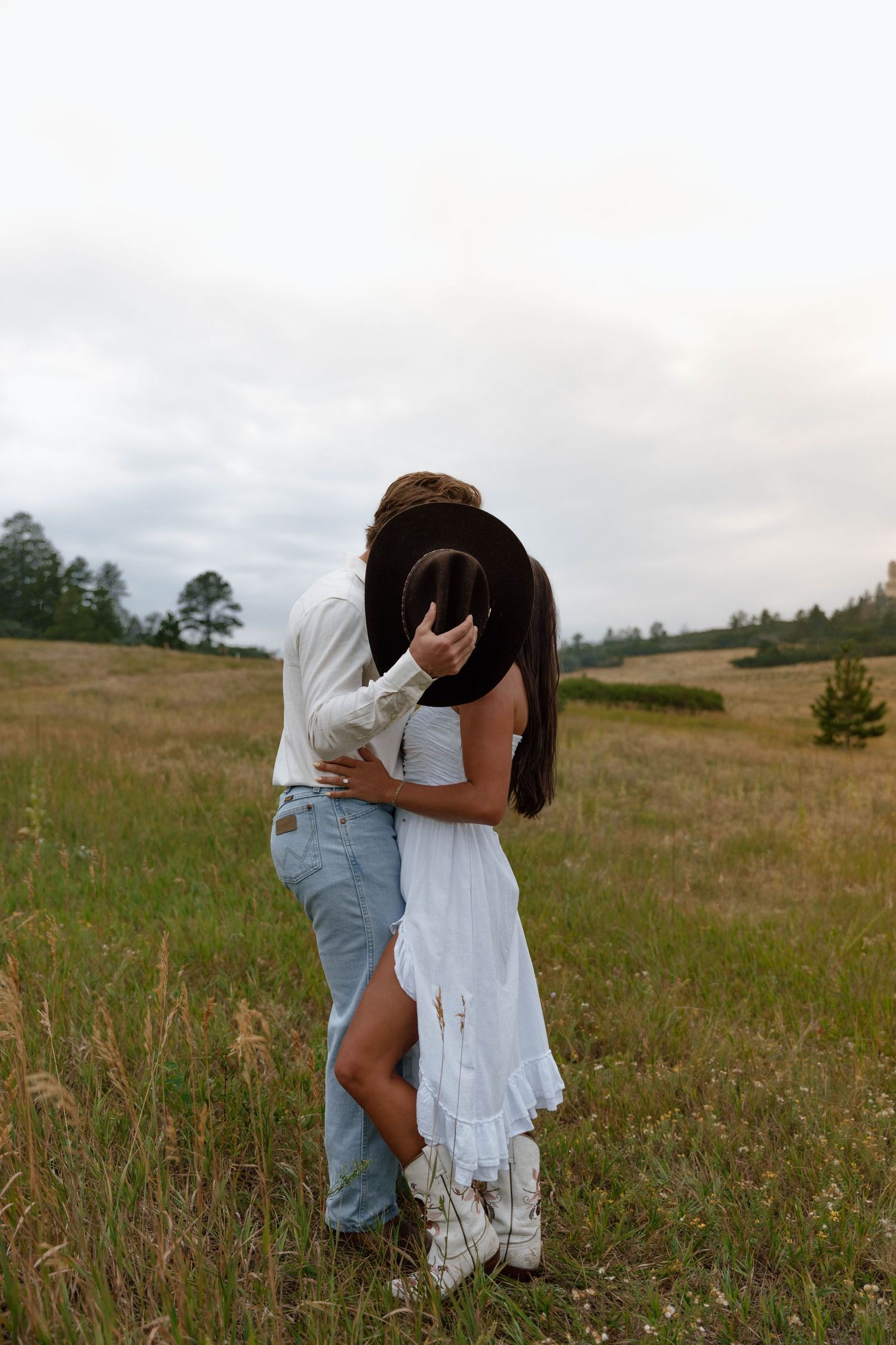 A romantic embrace in a rustic field with a white dress and denim against a cloudy mountain backdrop.