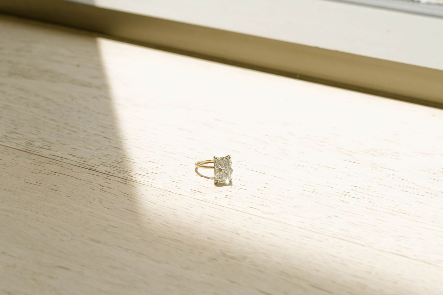 A diamond engagement ring casting a shadow on a white wooden surface in natural light.