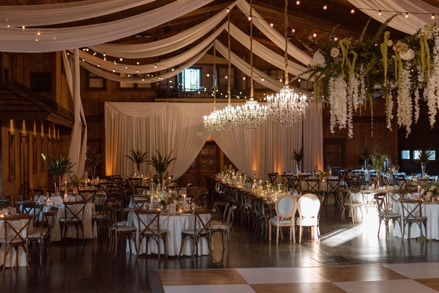 Stunning barn wedding venue with draped fabric ceiling, string lights, floral chandeliers, and long reception tables.
