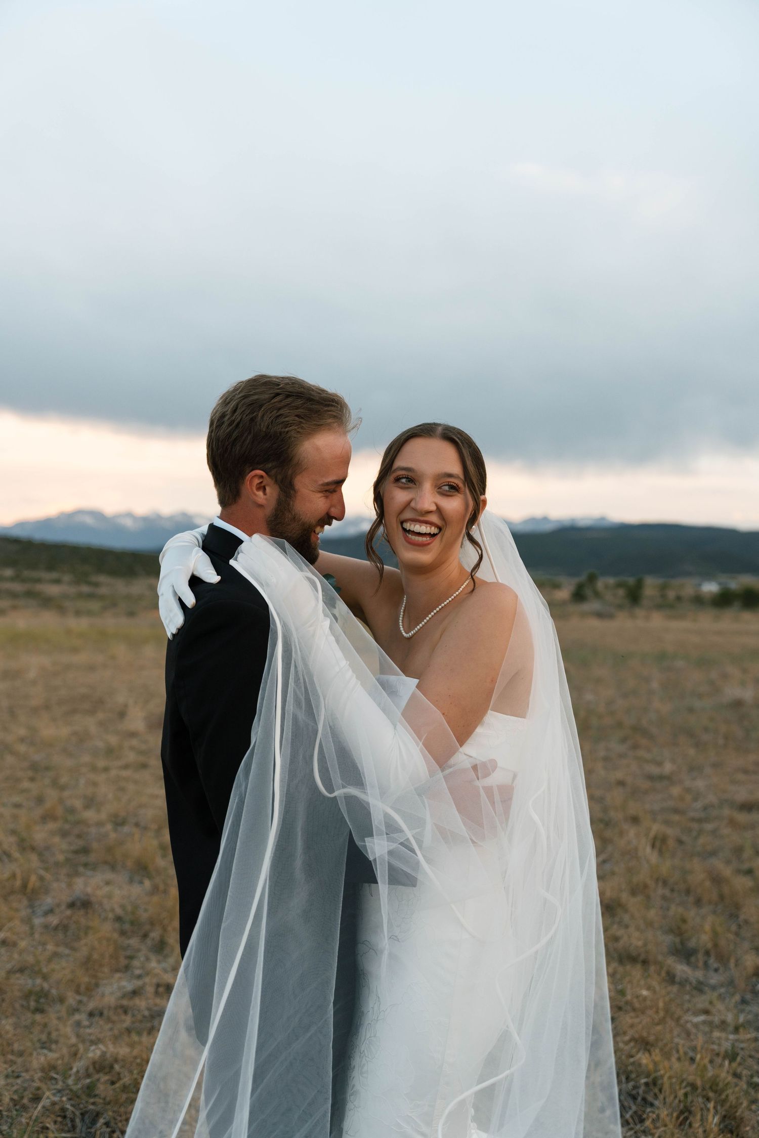 Happy bride and groom sharing a romantic moment in an open field at dusk with mountain views.