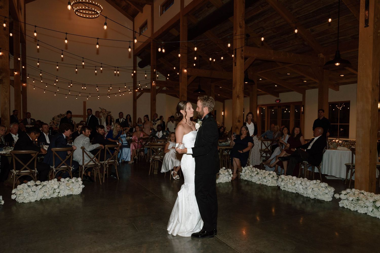Bride and groom share first dance in elegant barn reception hall with string lights and floral aisle.