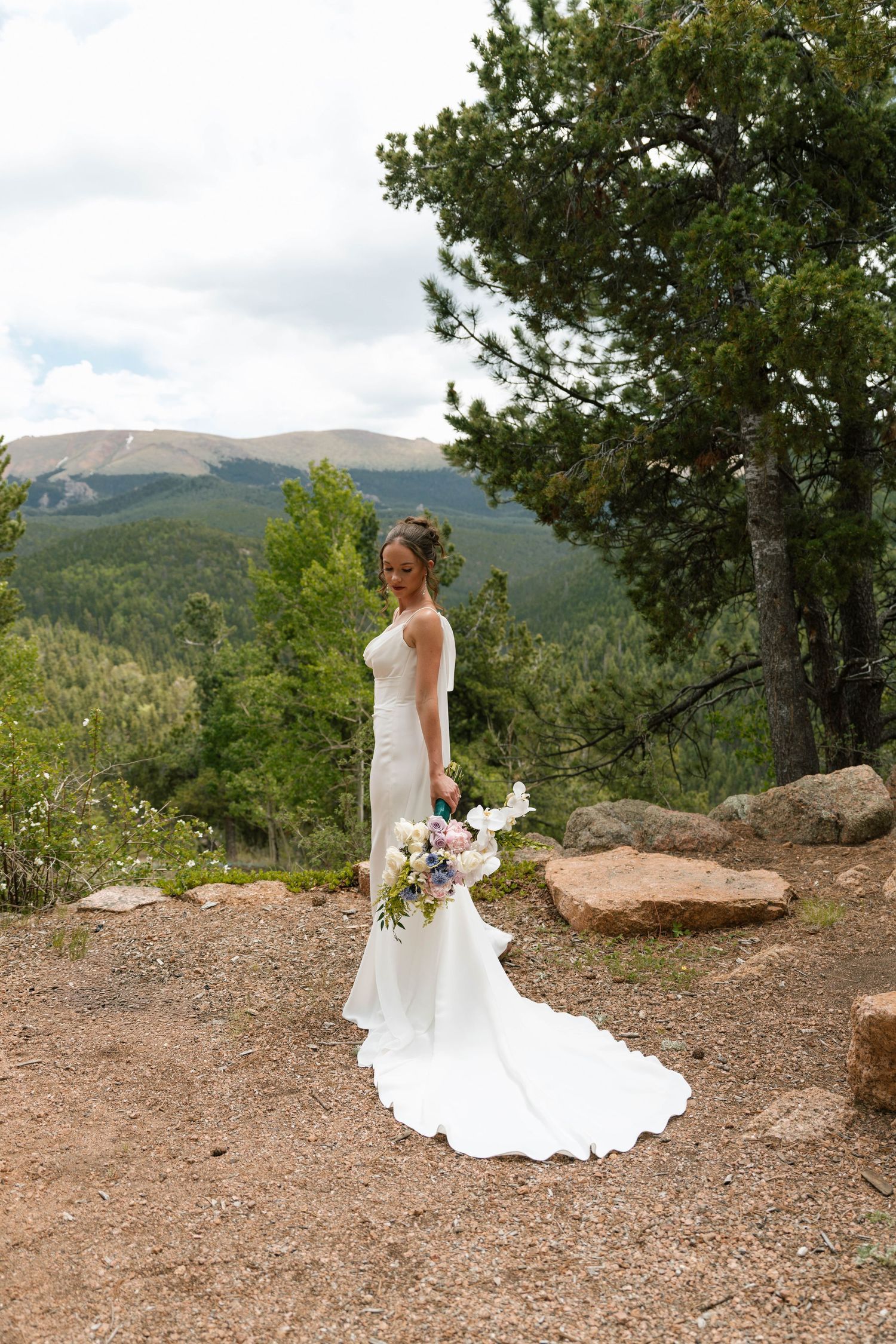 Bride in white gown holding bouquet with mountain forest backdrop on wedding day.