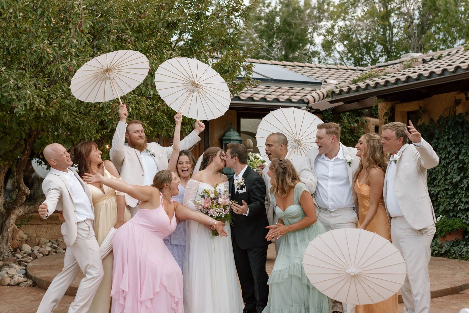Wedding party celebrates outdoors holding white parasols as bride and groom kiss, surrounded by guests in pastel attire.