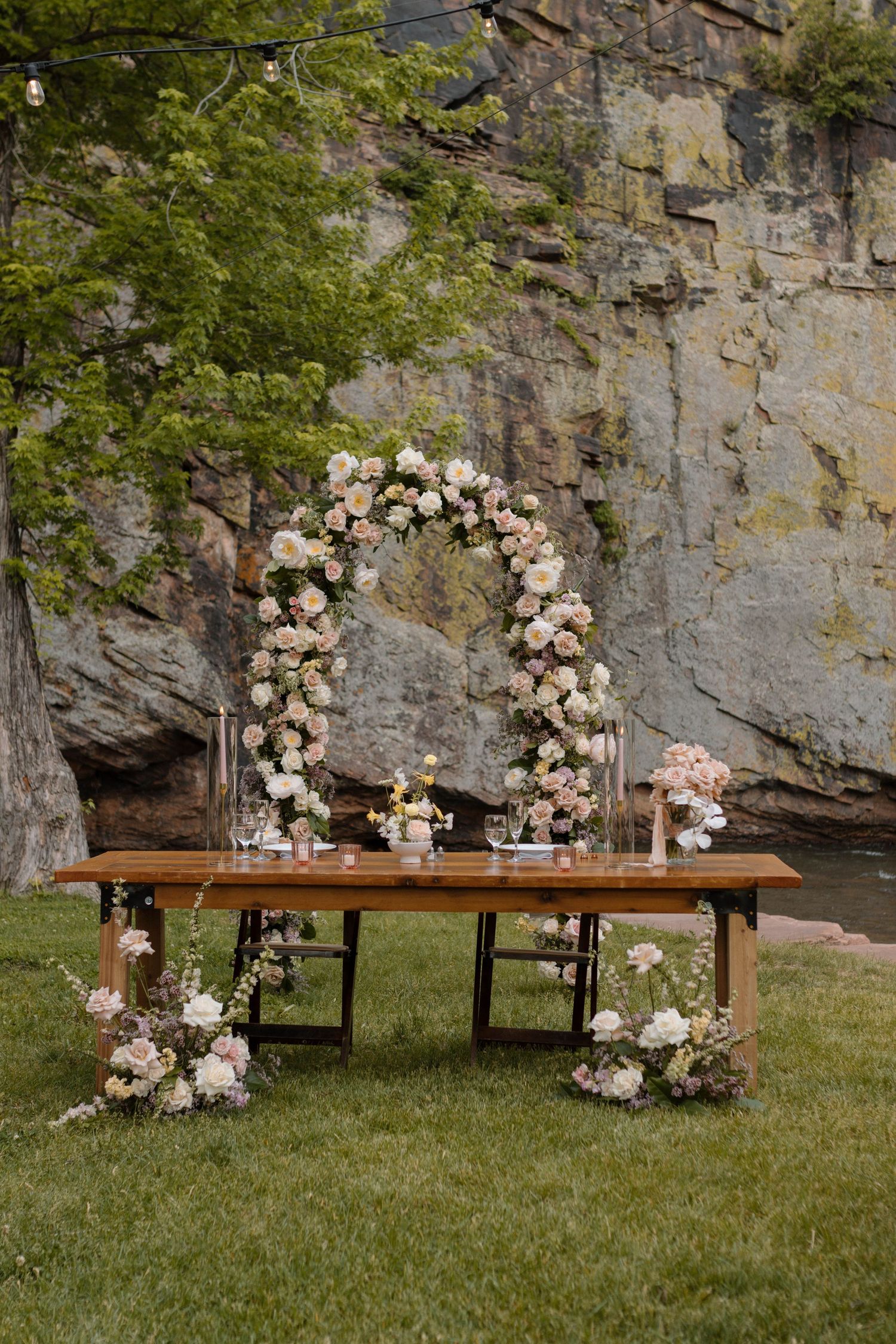 Outdoor wedding sweetheart table with floral arch, blush roses, wood farm table, and rocky cliff backdrop on green lawn.
