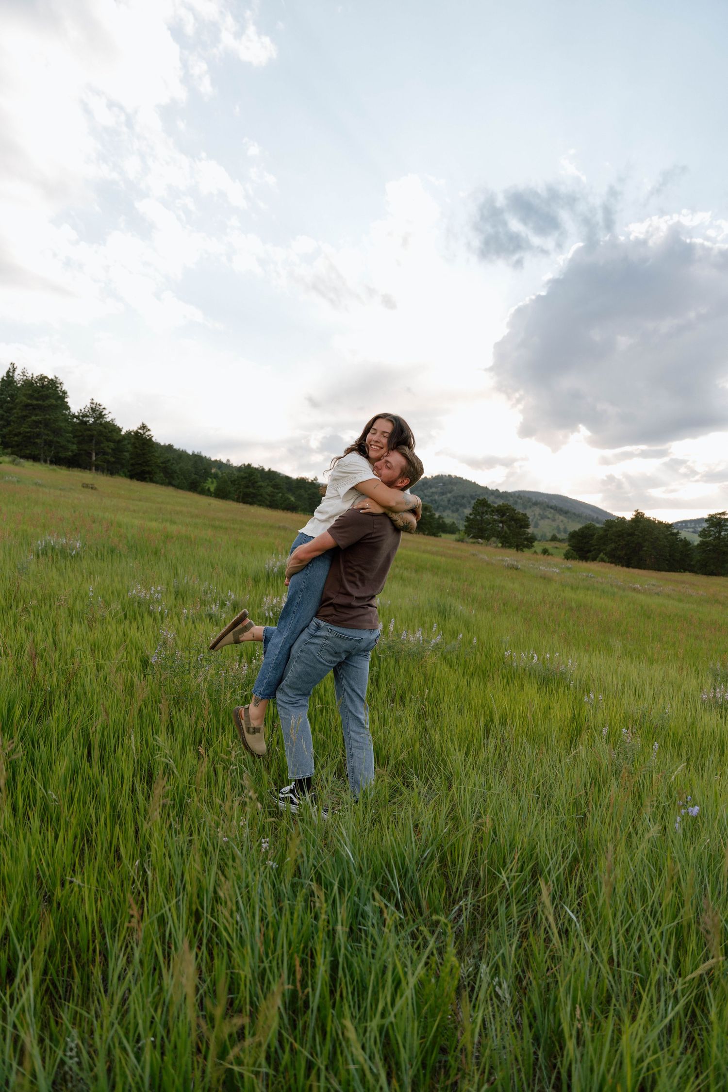 Two people playing and embracing in tall grass during golden hour with mountains in the distance.