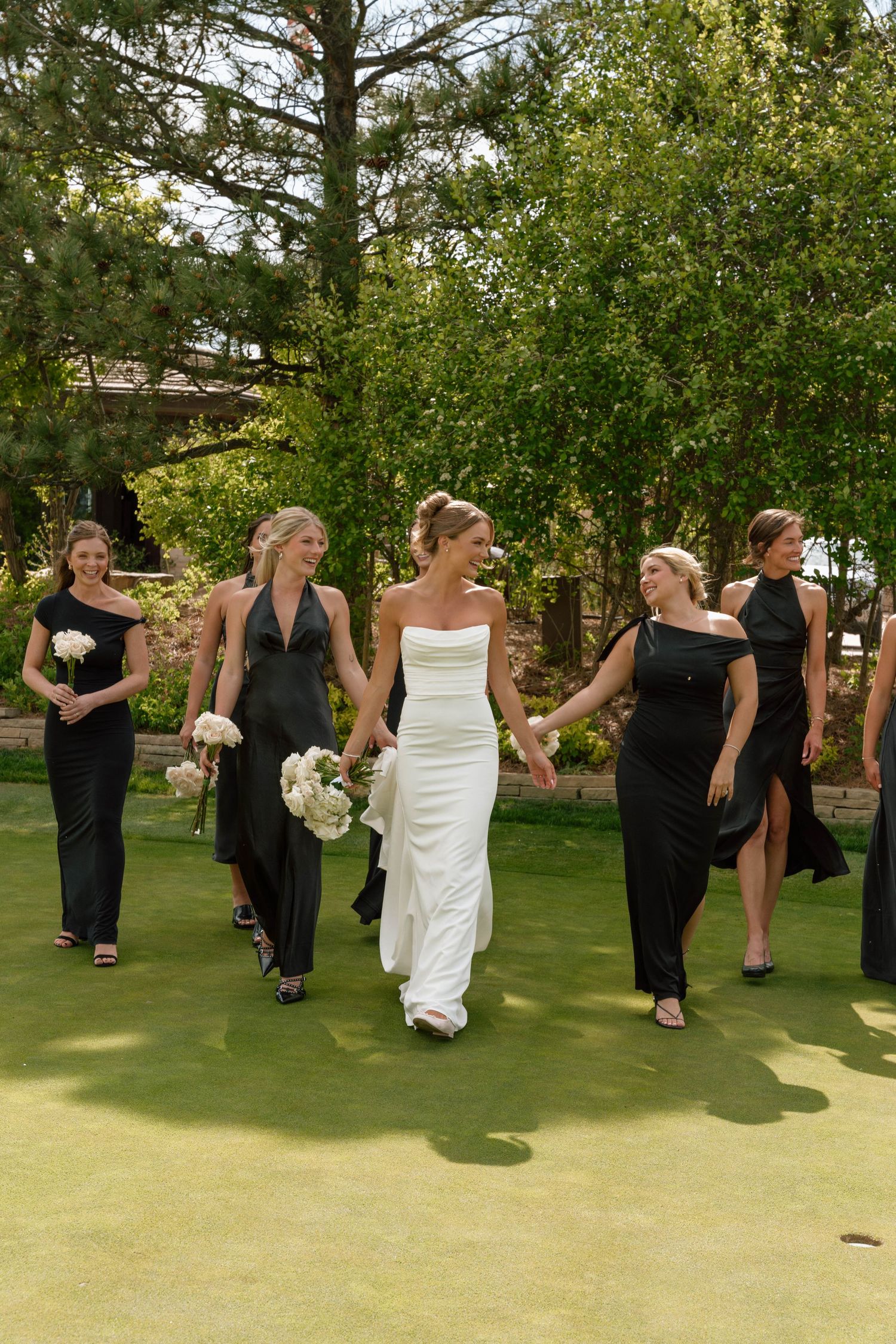 A wedding party walking together on a green lawn with trees in the background.