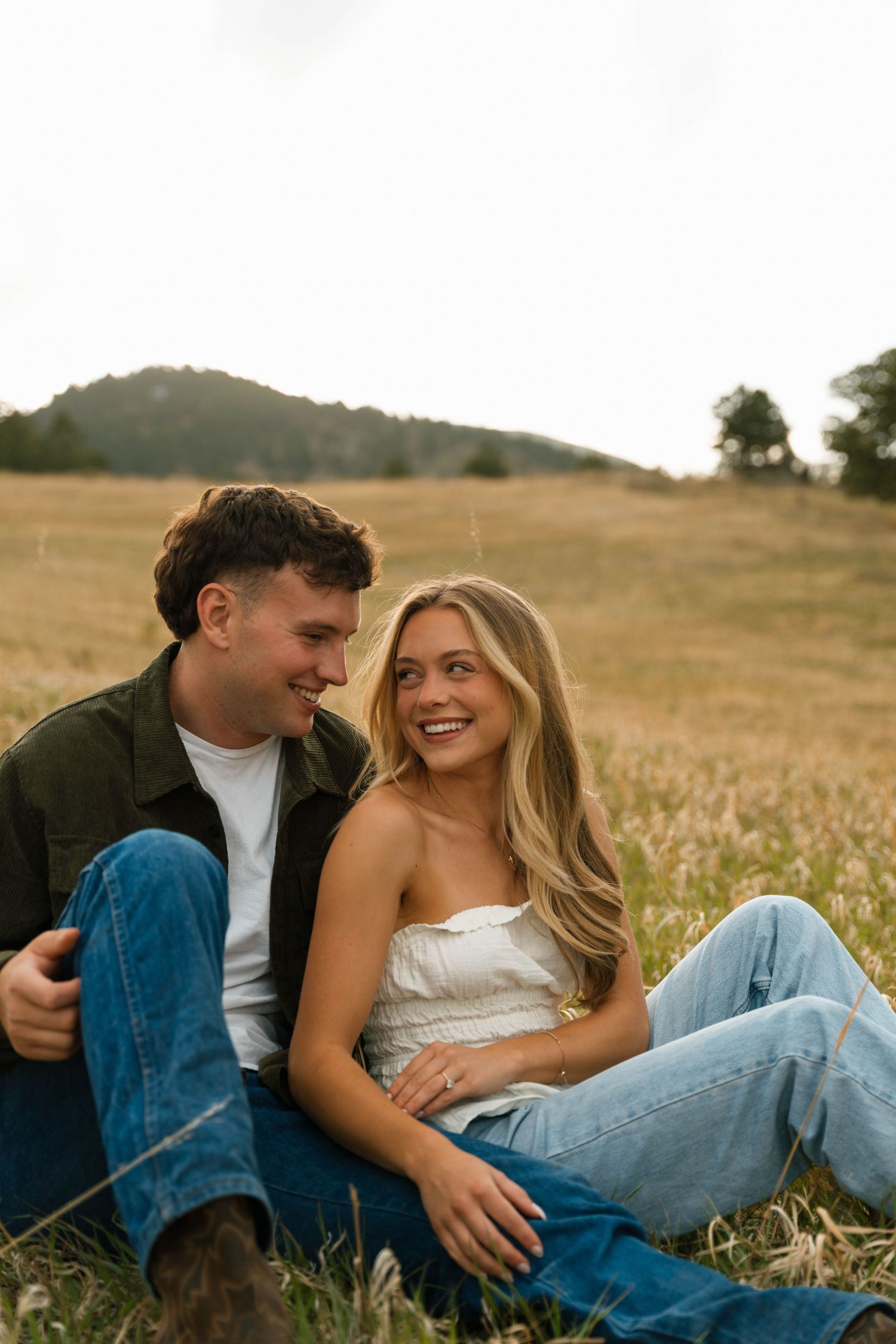 Couple sitting together in a golden field, smiling and laughing, woman in white top and jeans, man in green jacket.