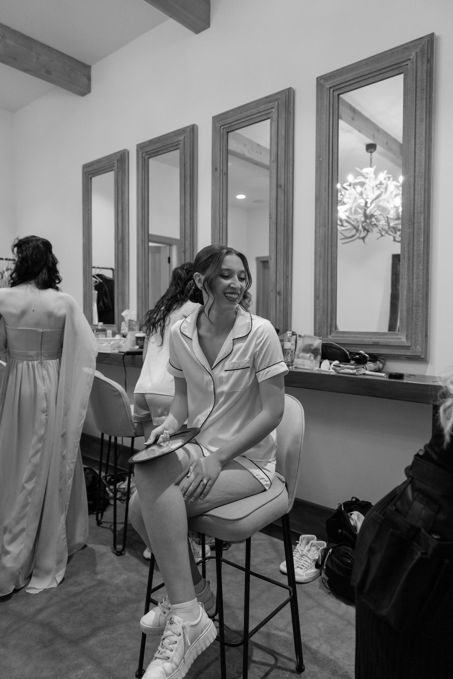Bride laughing in pajamas on a stool in a bridal suite with mirrors and bridesmaids getting ready.