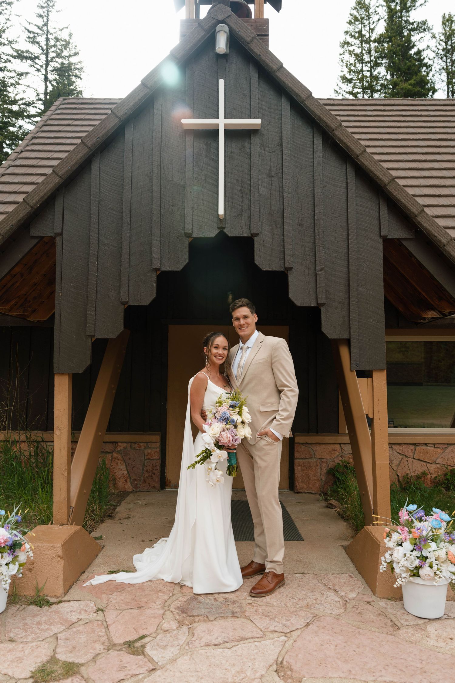 Bride and groom pose outside a small dark wood chapel with a white cross after their wedding ceremony.