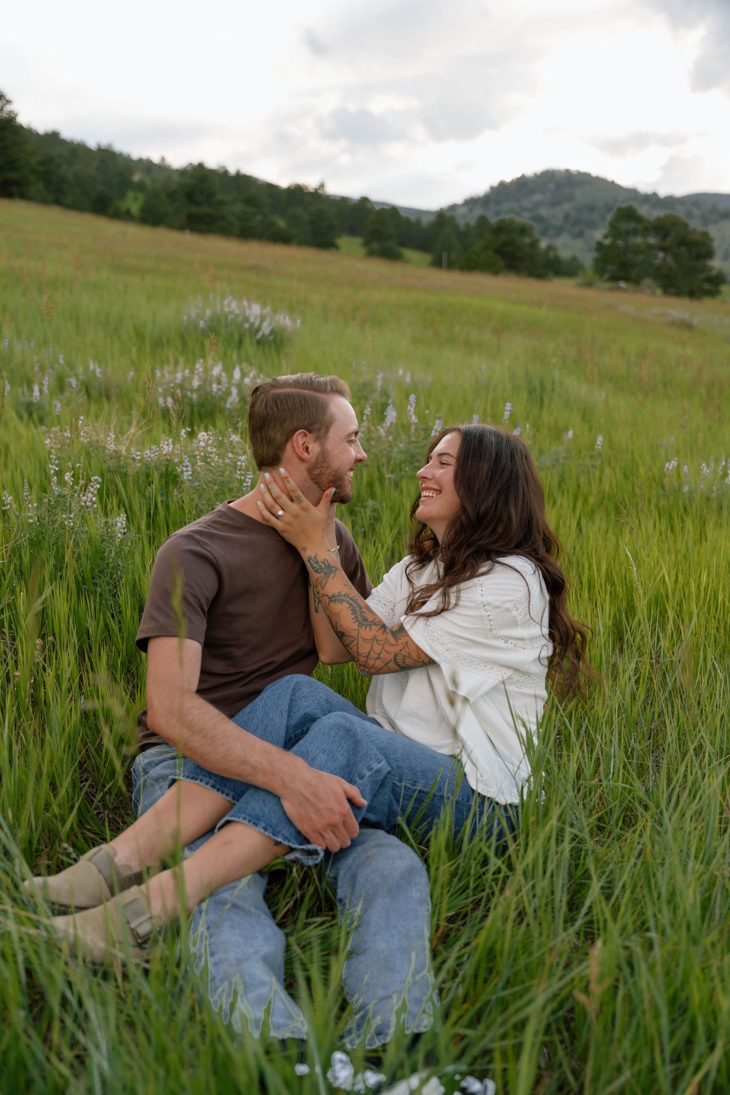 A couple shares a tender moment while sitting in a wildflower meadow with mountains in the background.