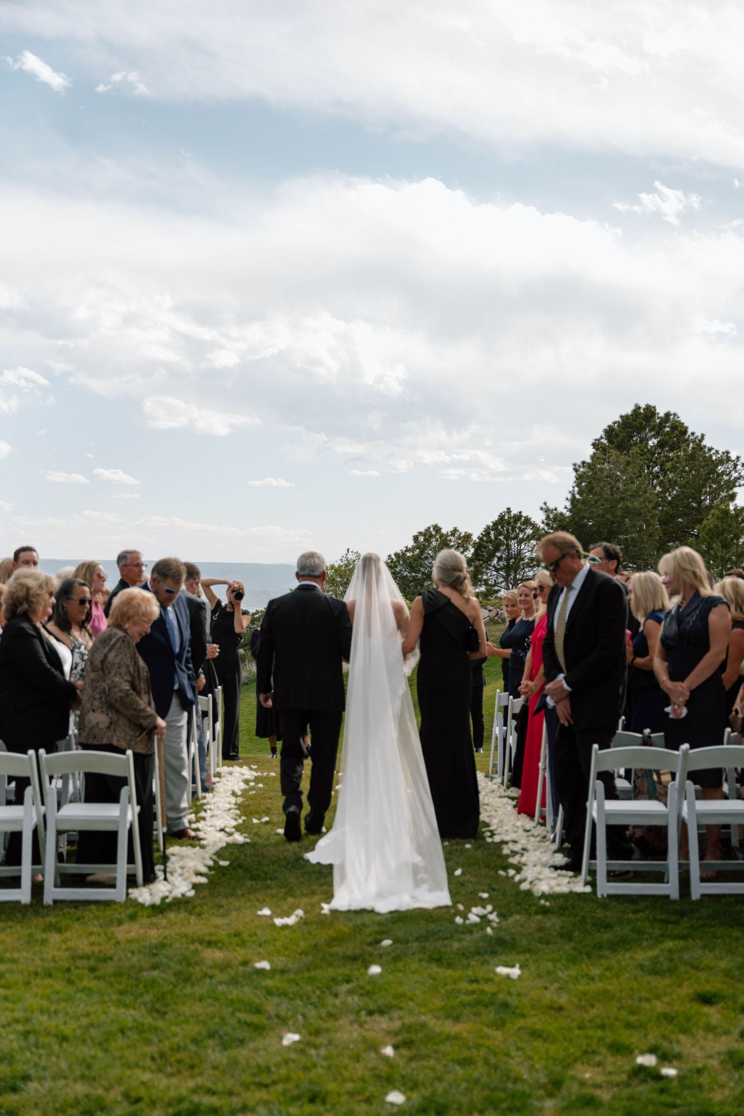Outdoor wedding processional with white chairs and flower petals lining the aisle.