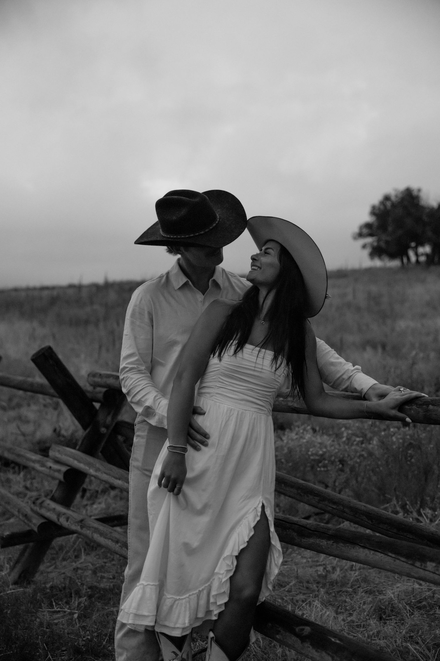A romantic black and white portrait by a wooden fence featuring cowboy hats and a flowing white dress.