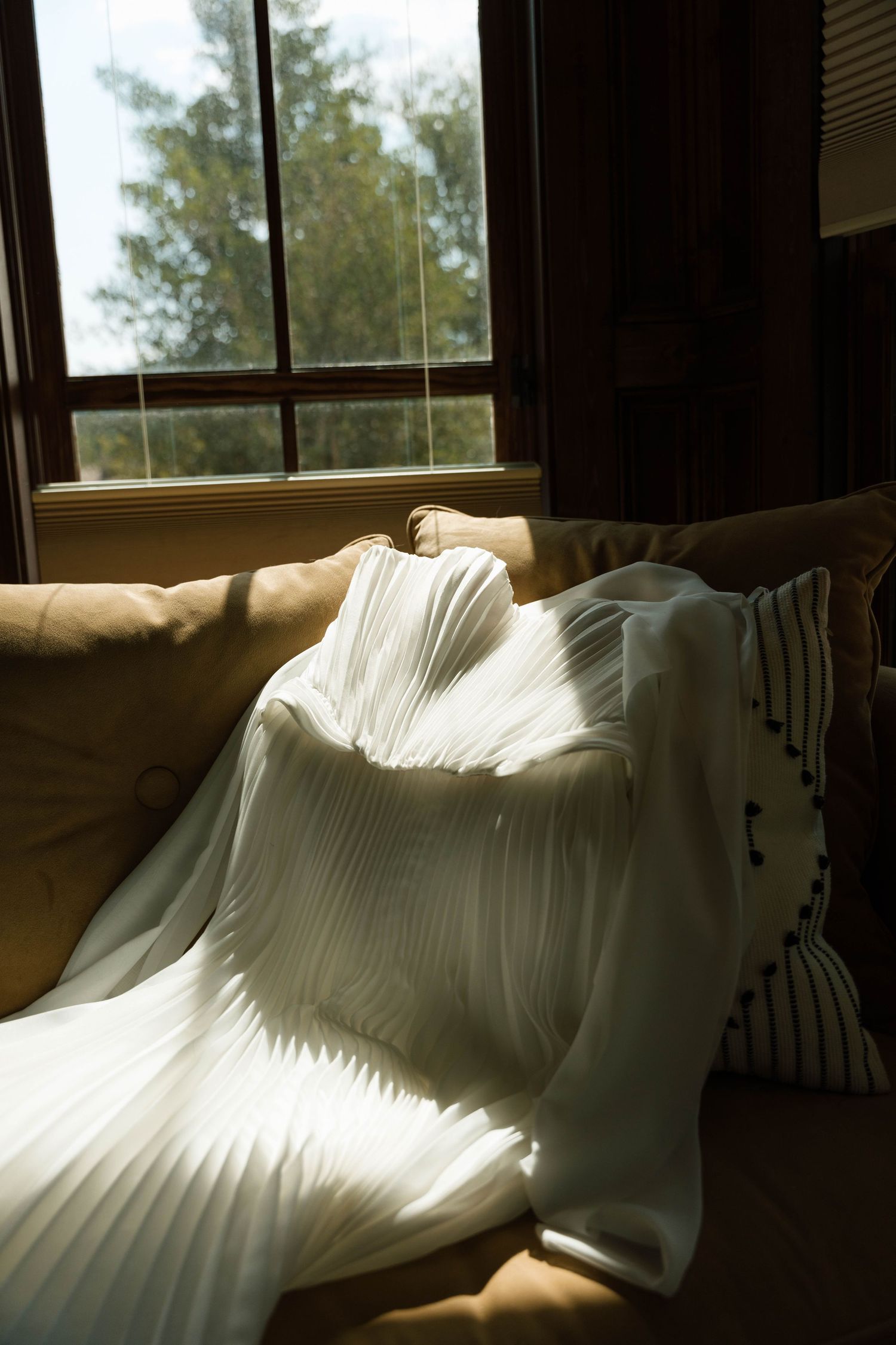 White pleated fabric draped over a sunlit sofa near a wooden window with trees visible outside.