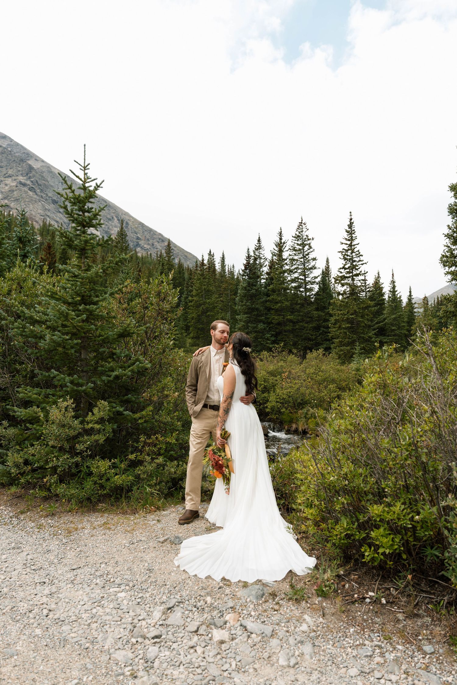 Bride and groom share a romantic kiss surrounded by evergreen trees and mountains in a stunning outdoor wedding setting.