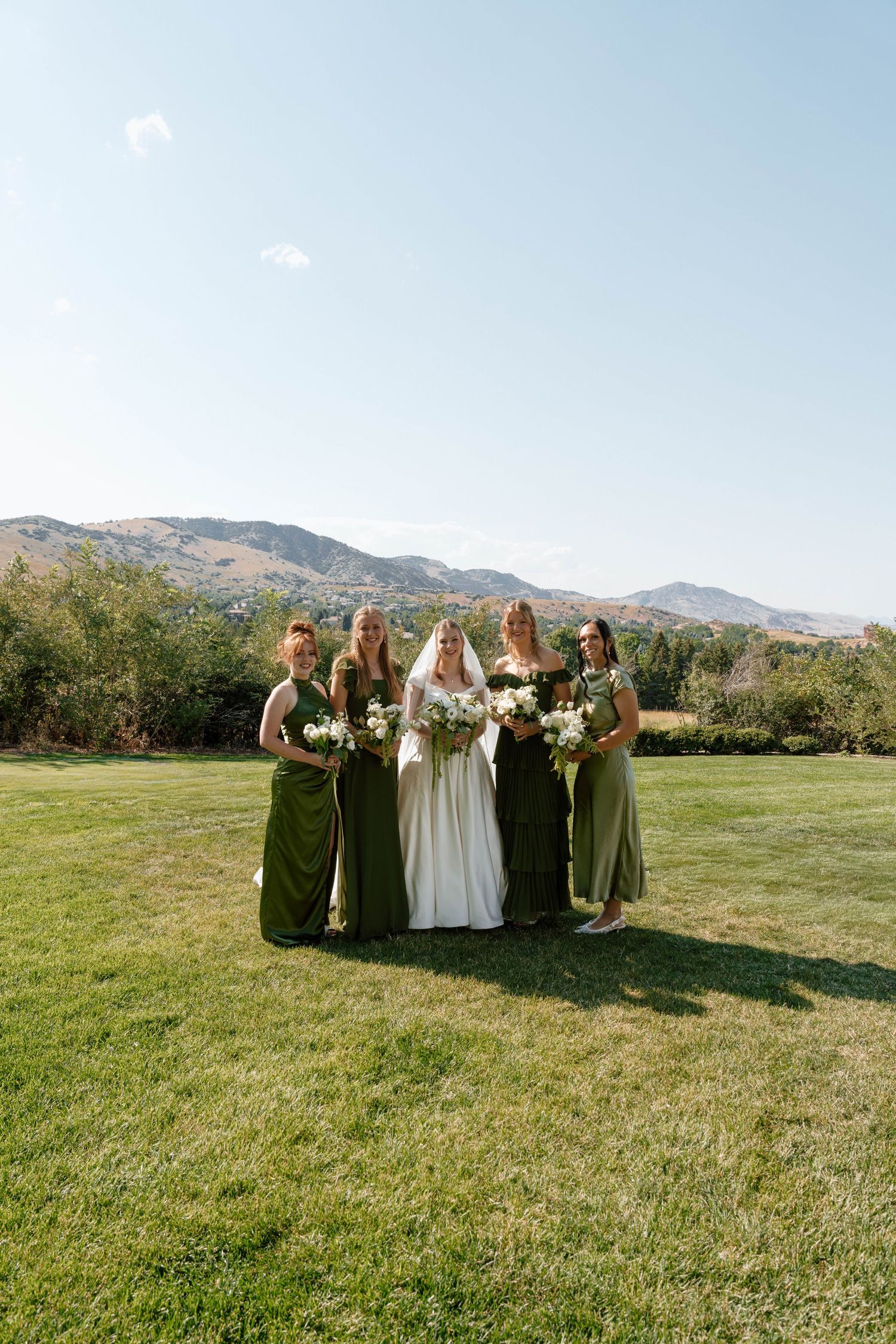 Wedding party in olive green dresses poses on lawn with mountains in background.