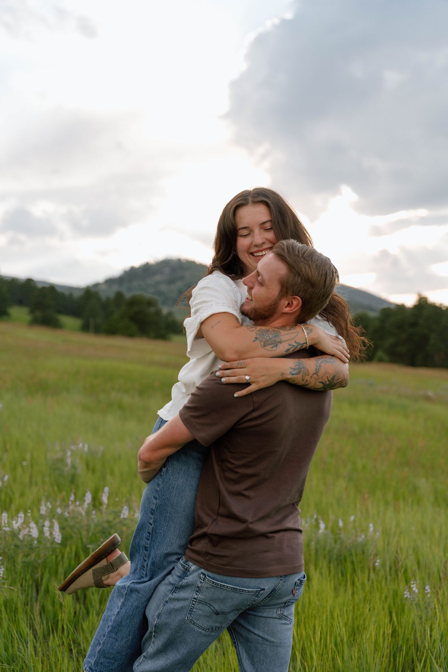 A romantic embrace captured in a sunlit mountain meadow with tall grass and cloudy skies.