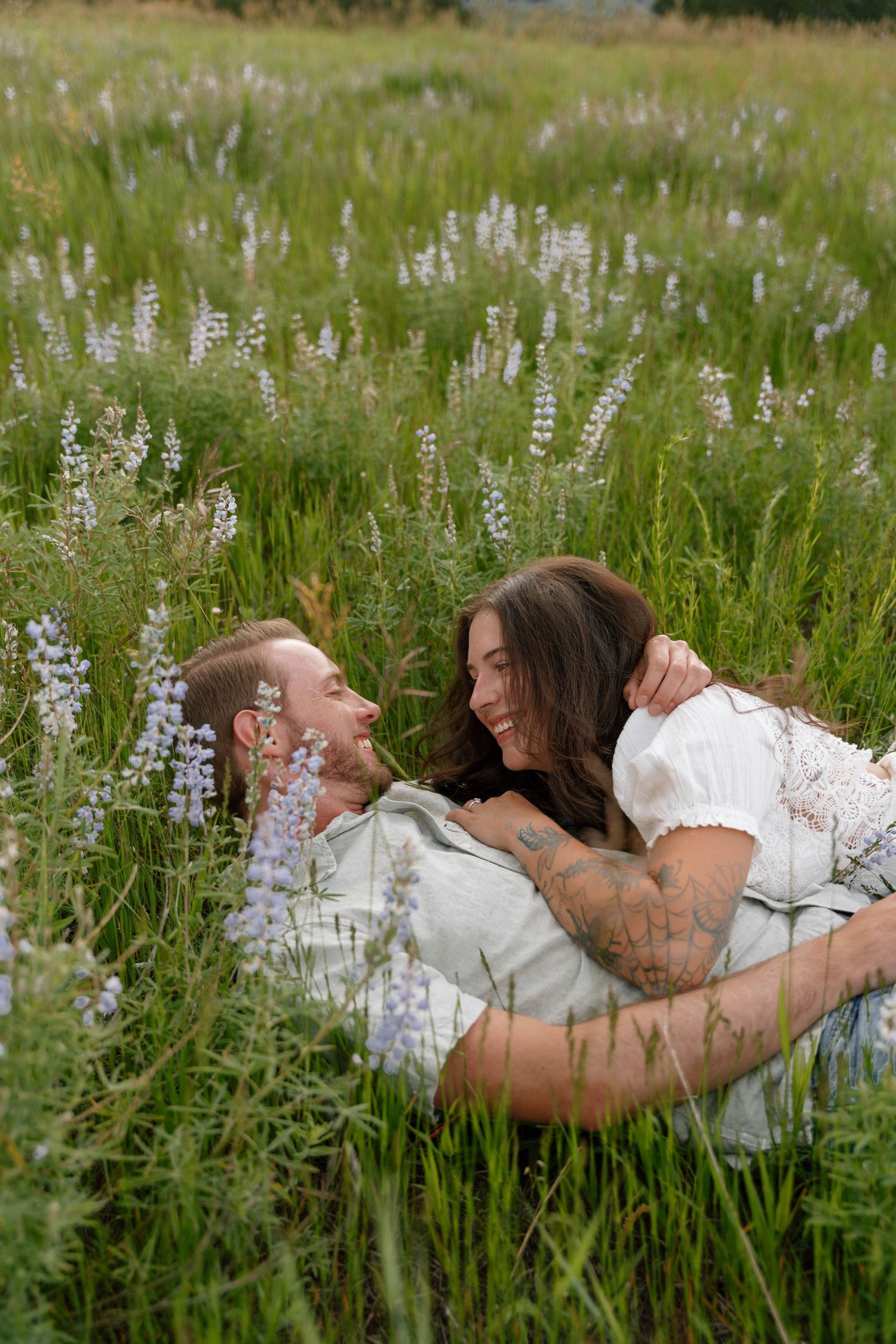 Couple laying in tall grass surrounded by white and purple wildflowers on a sunny day.
