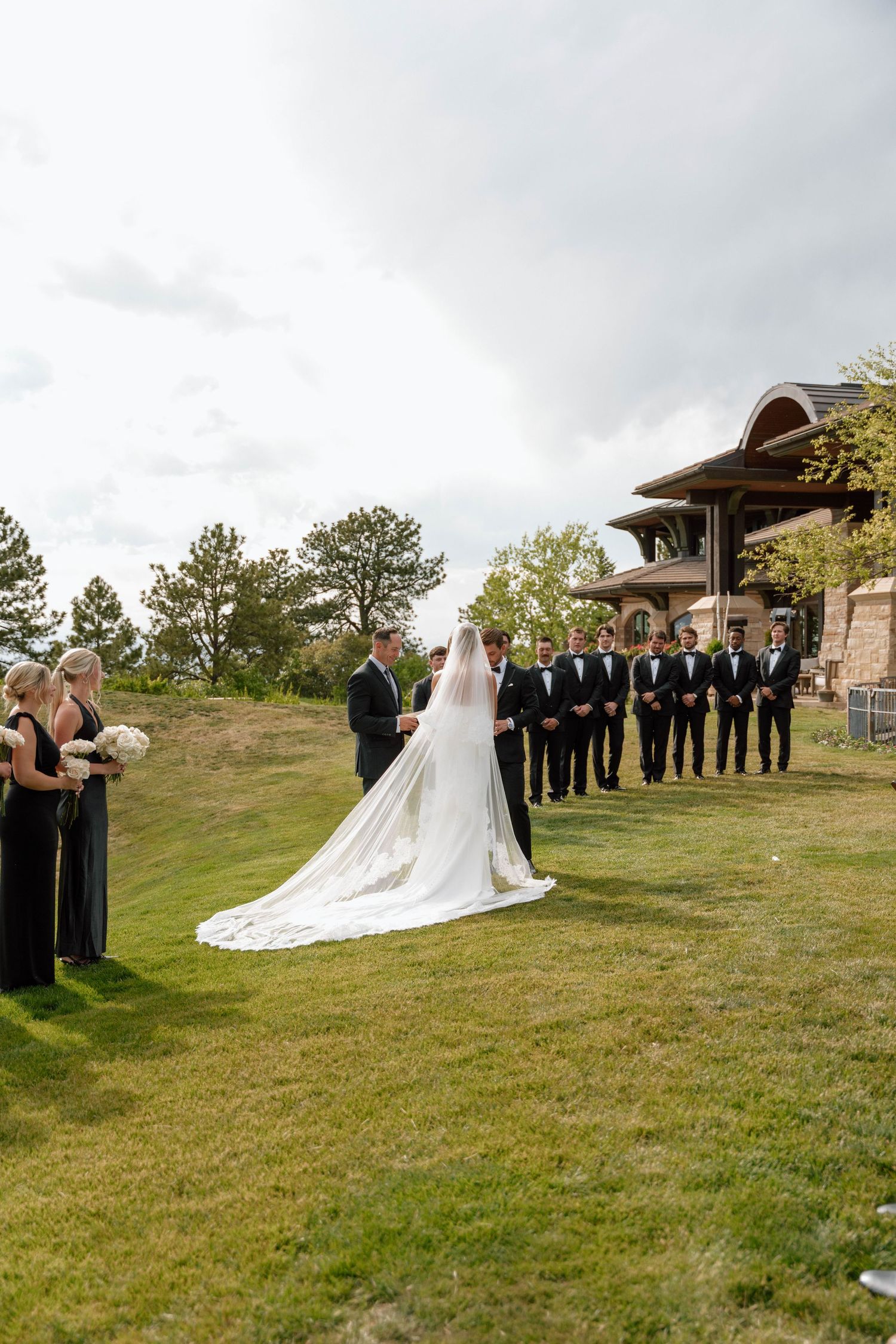 Wedding ceremony on scenic hillside with wedding party in black attire and flowing white veil.