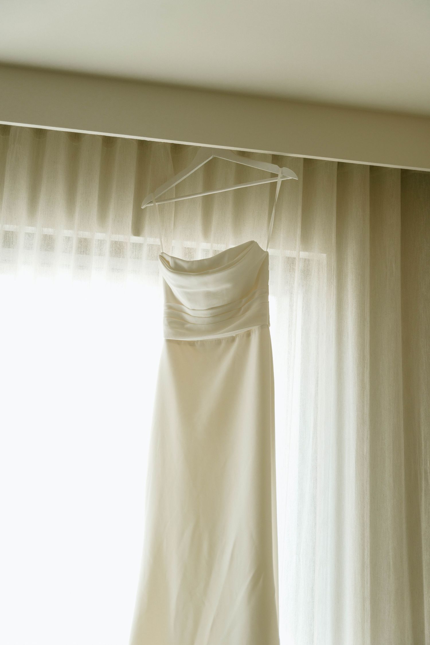 A white wedding dress hangs elegantly against sheer curtains in soft natural light.