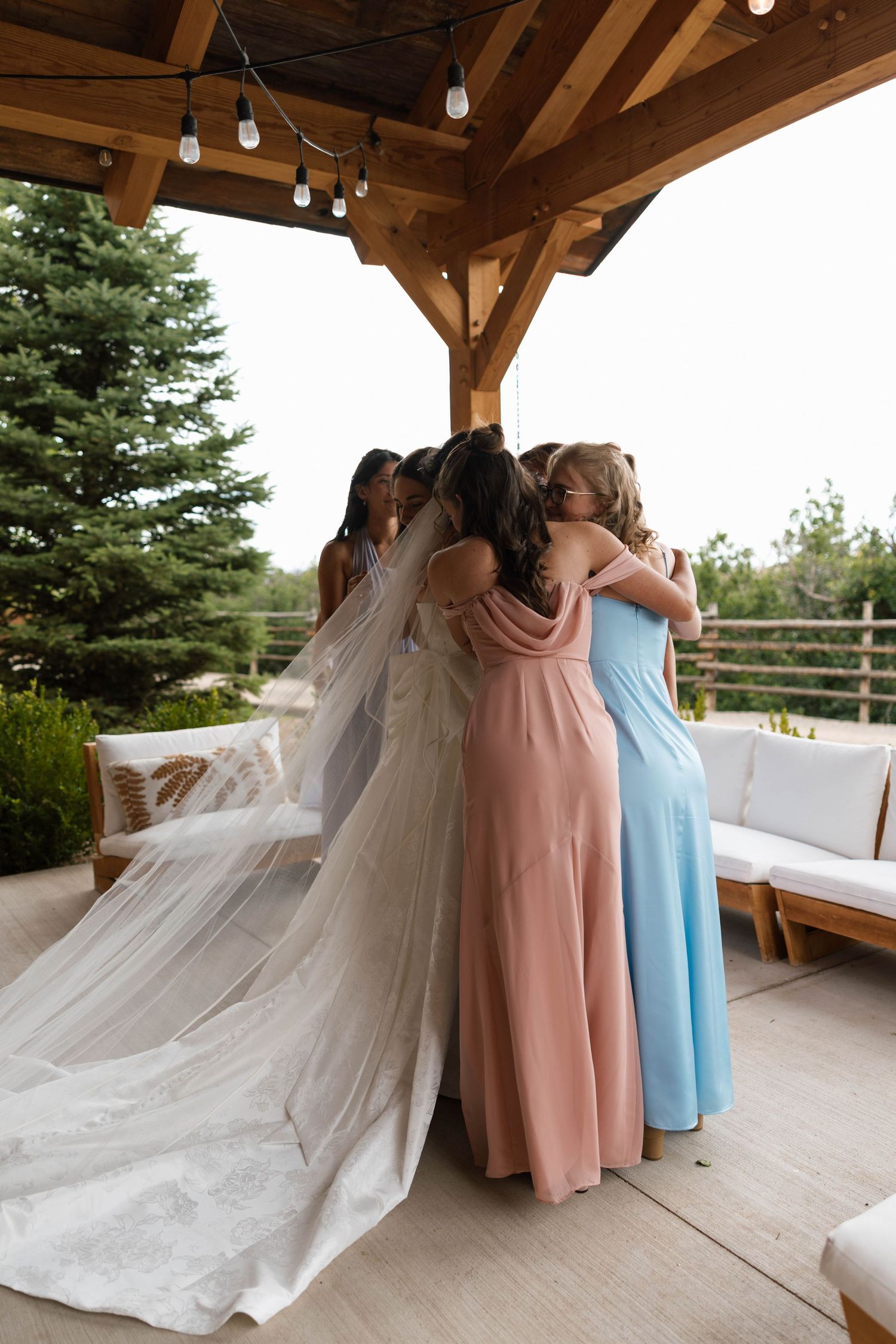 Bride in white gown and veil sharing a group hug with bridesmaids in pink and blue dresses outdoors.