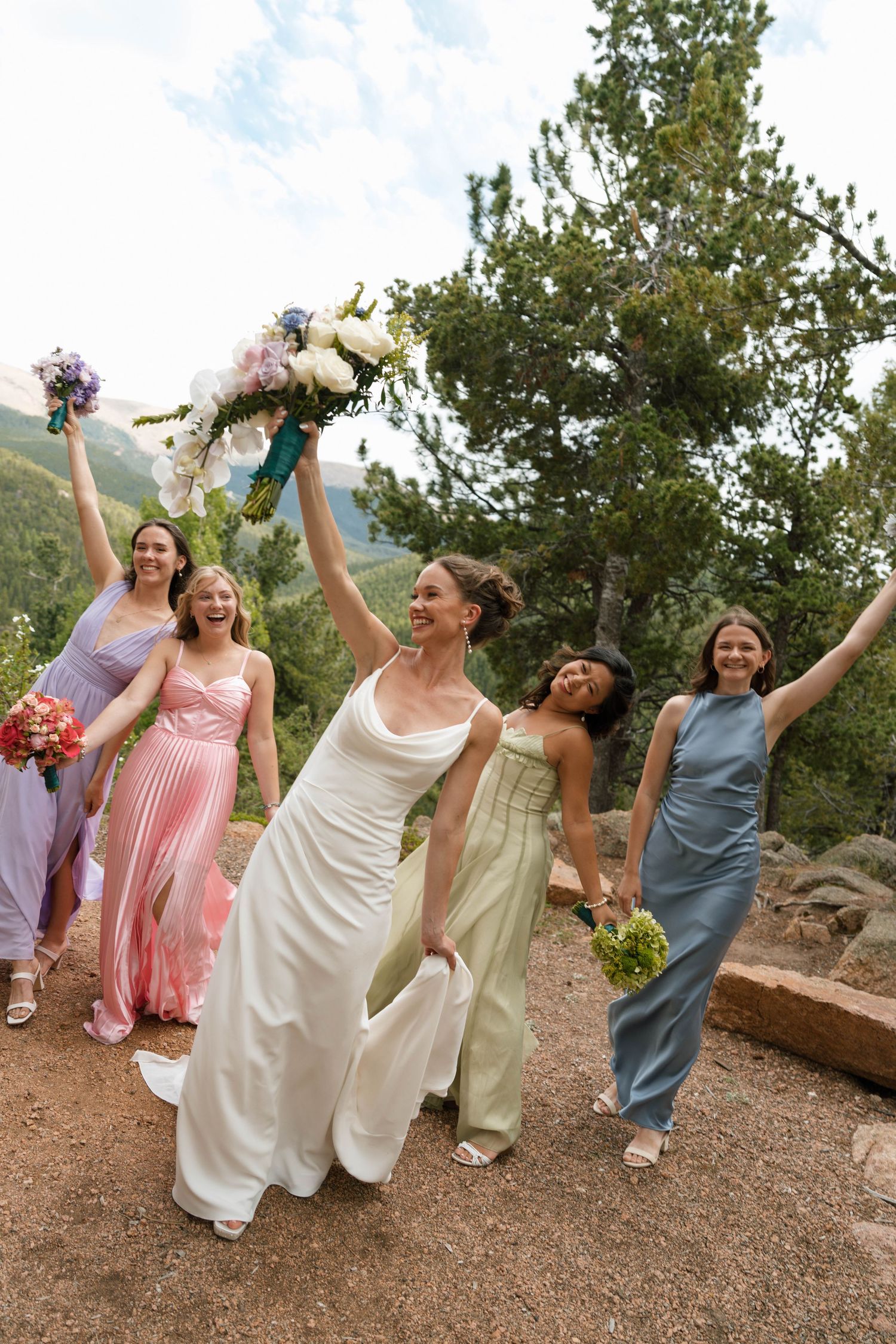 Bride and bridesmaids in colorful gowns celebrating outdoors with raised bouquets.